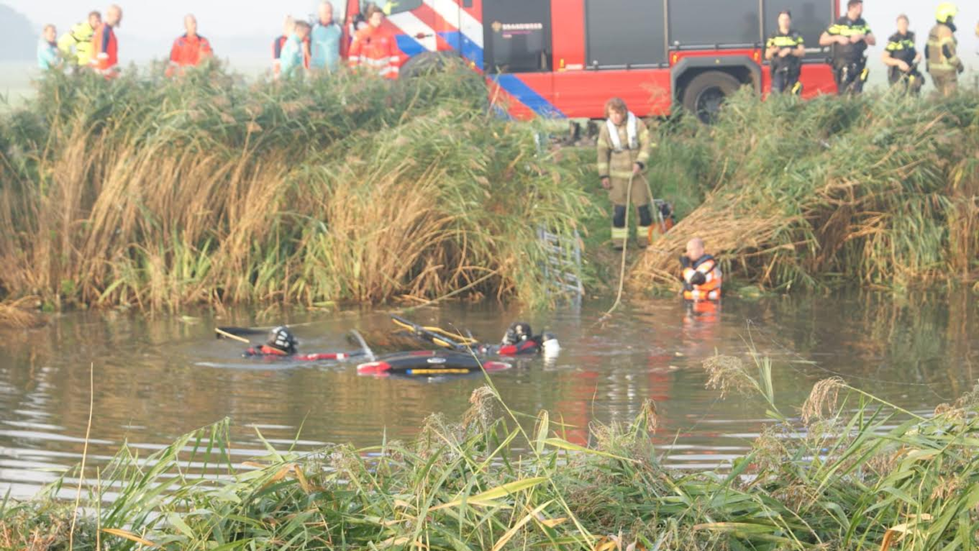 Brandweermannen en duikers onderzoeken een sloot, met een brandweerwagen en andere hulpverleners op de achtergrond.