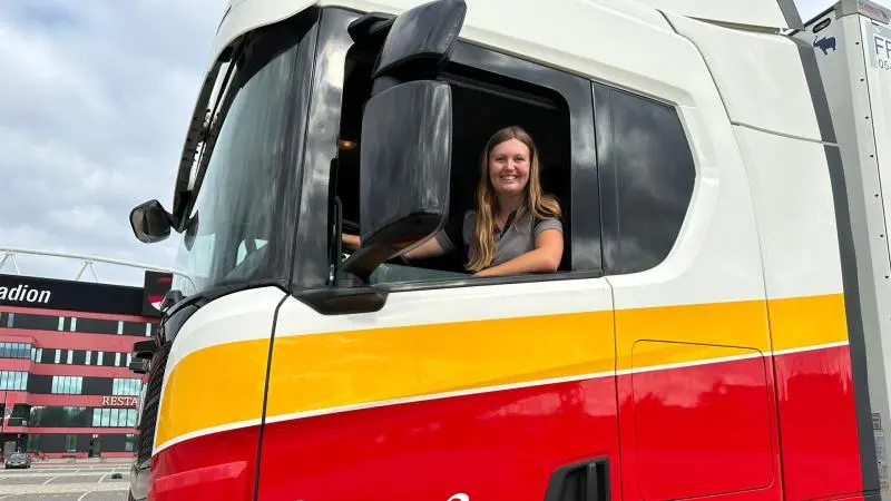 Vrouw glimlacht terwijl ze achter het stuur van een vrachtwagen zit.