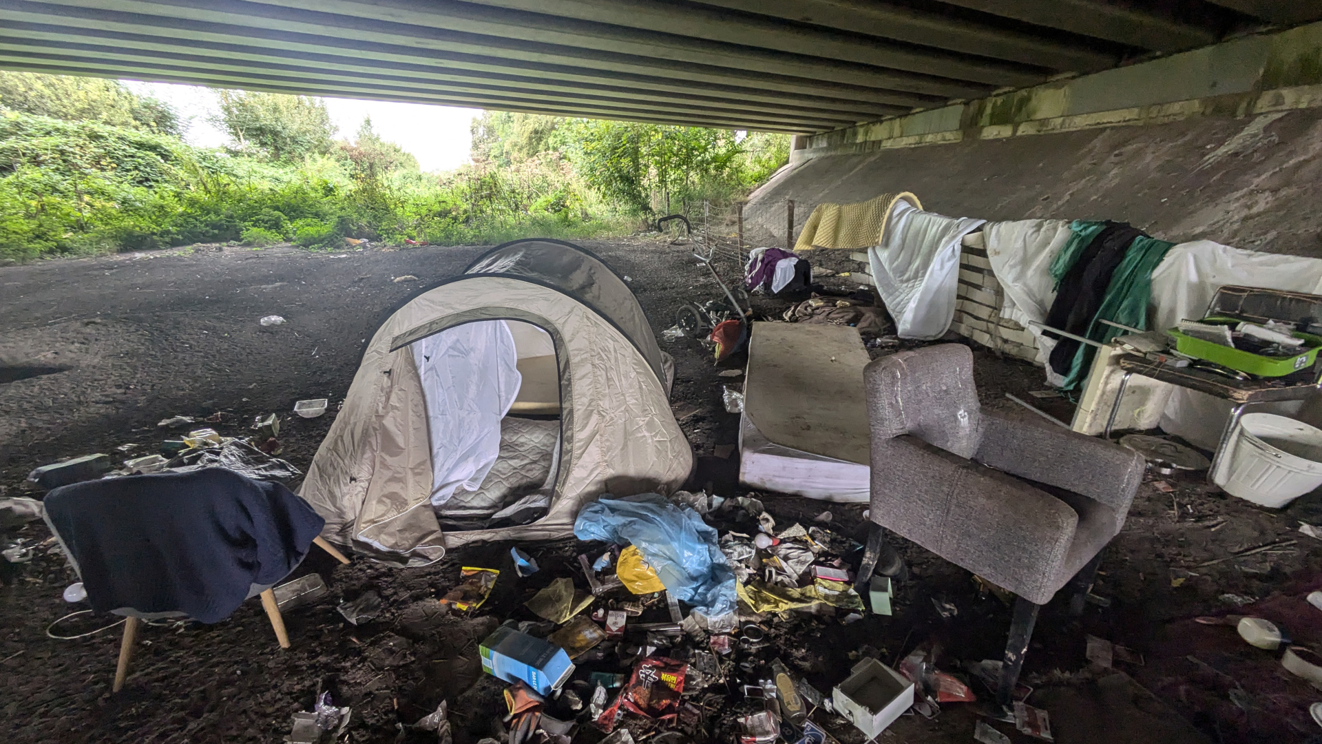 Kampeerplek onder een brug met een tent, stoelen, matrassen en verspreide rommel.