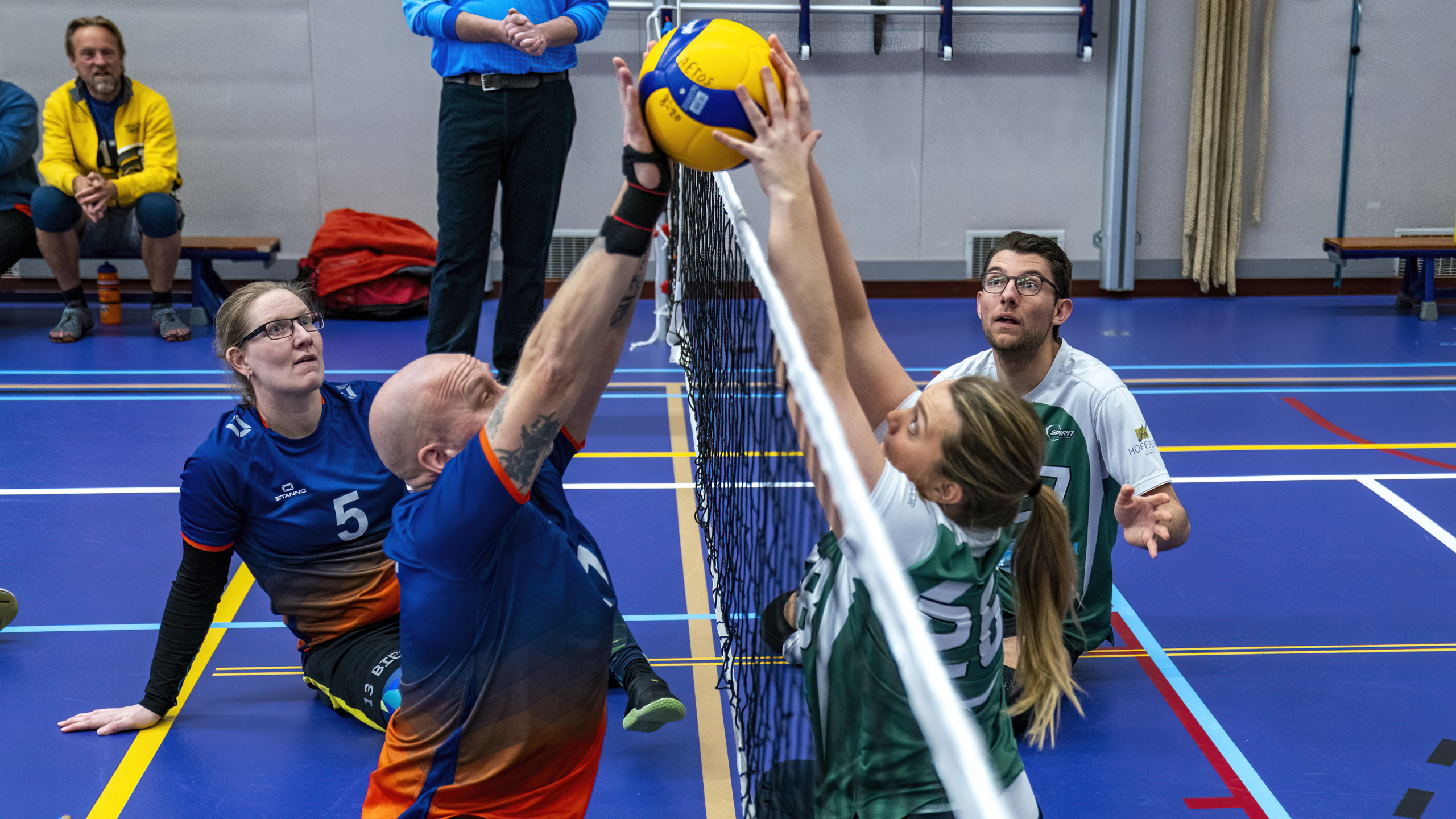 Zittend volleybalwedstrijd waarbij een man en een vrouw de bal boven het net blokkeren.