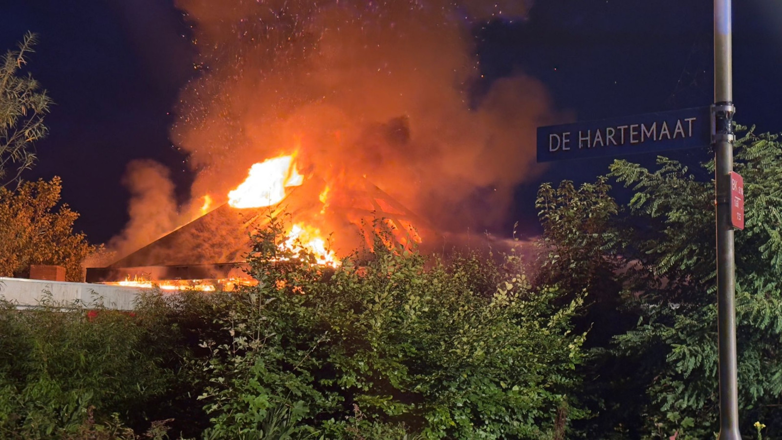 Brandend gebouw omgeven door bomen, met zichtbare straatnaambord "De Hartemaat" aan een lantaarnpaal.