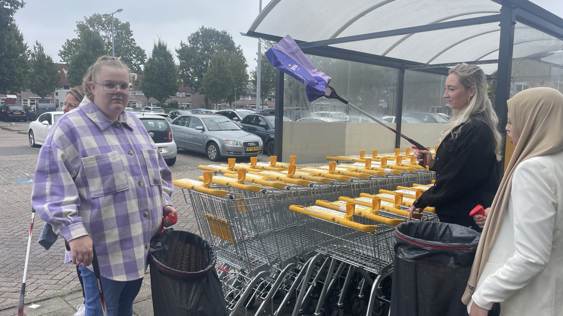 Drie mensen verzamelen afval met afvalgrijpers bij een parkeerplaats met winkelwagentjes.