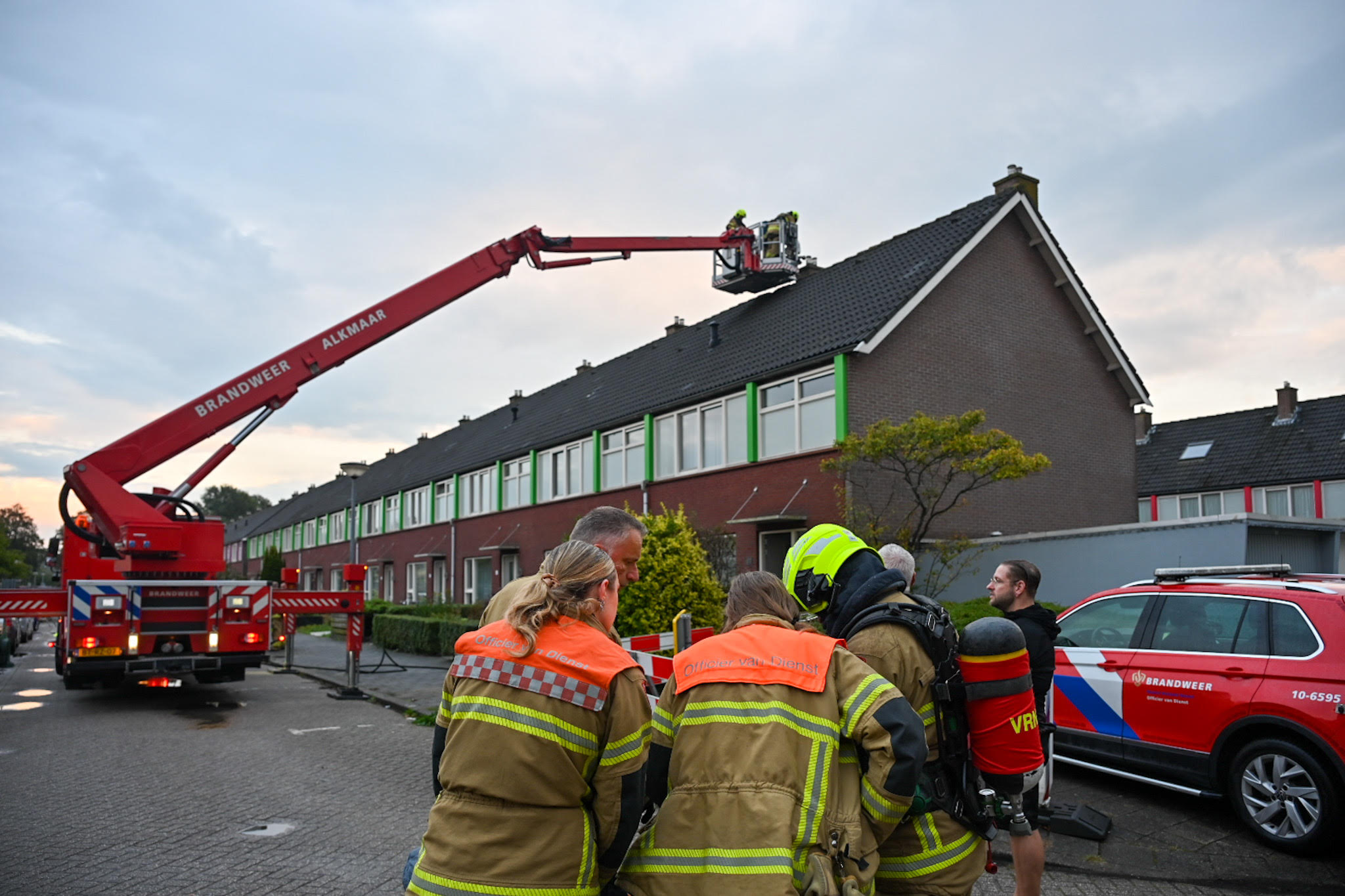 Brandweerlieden bij een brandweerwagen met een ladder die naar het dak van een rijtjeshuis reikt.