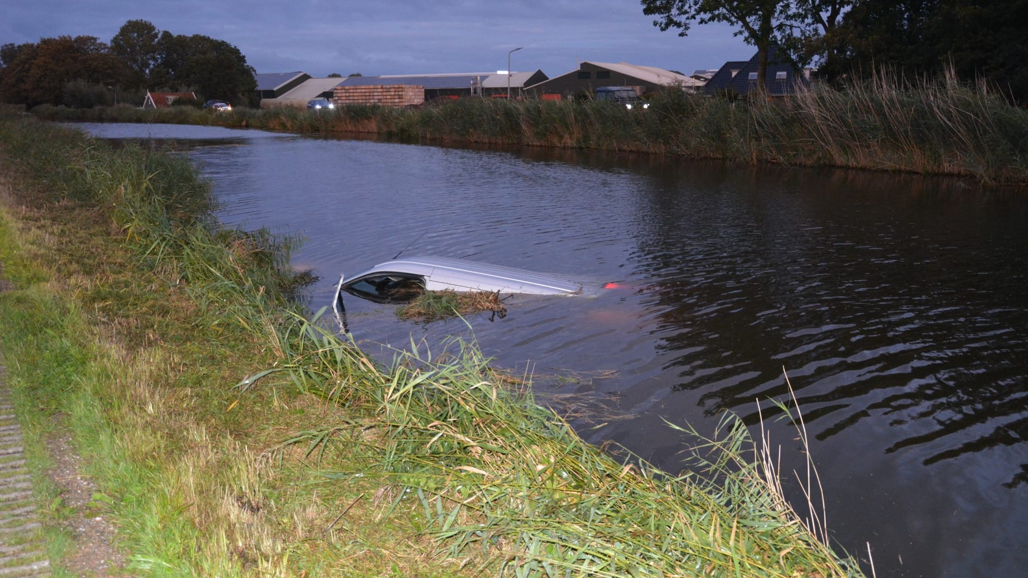 Auto deels onder water in kanaal naast landweg, met weiland en gebouwen op achtergrond.