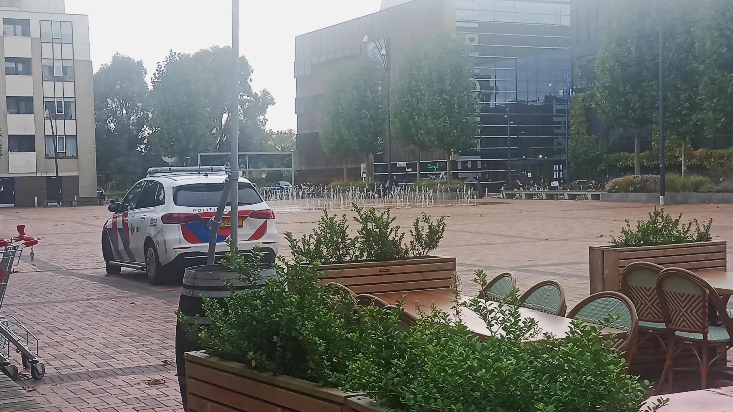 Een politiewagen geparkeerd op een plein met een fontein op de achtergrond en enkele gebouwen, waaronder een glazen gebouw.