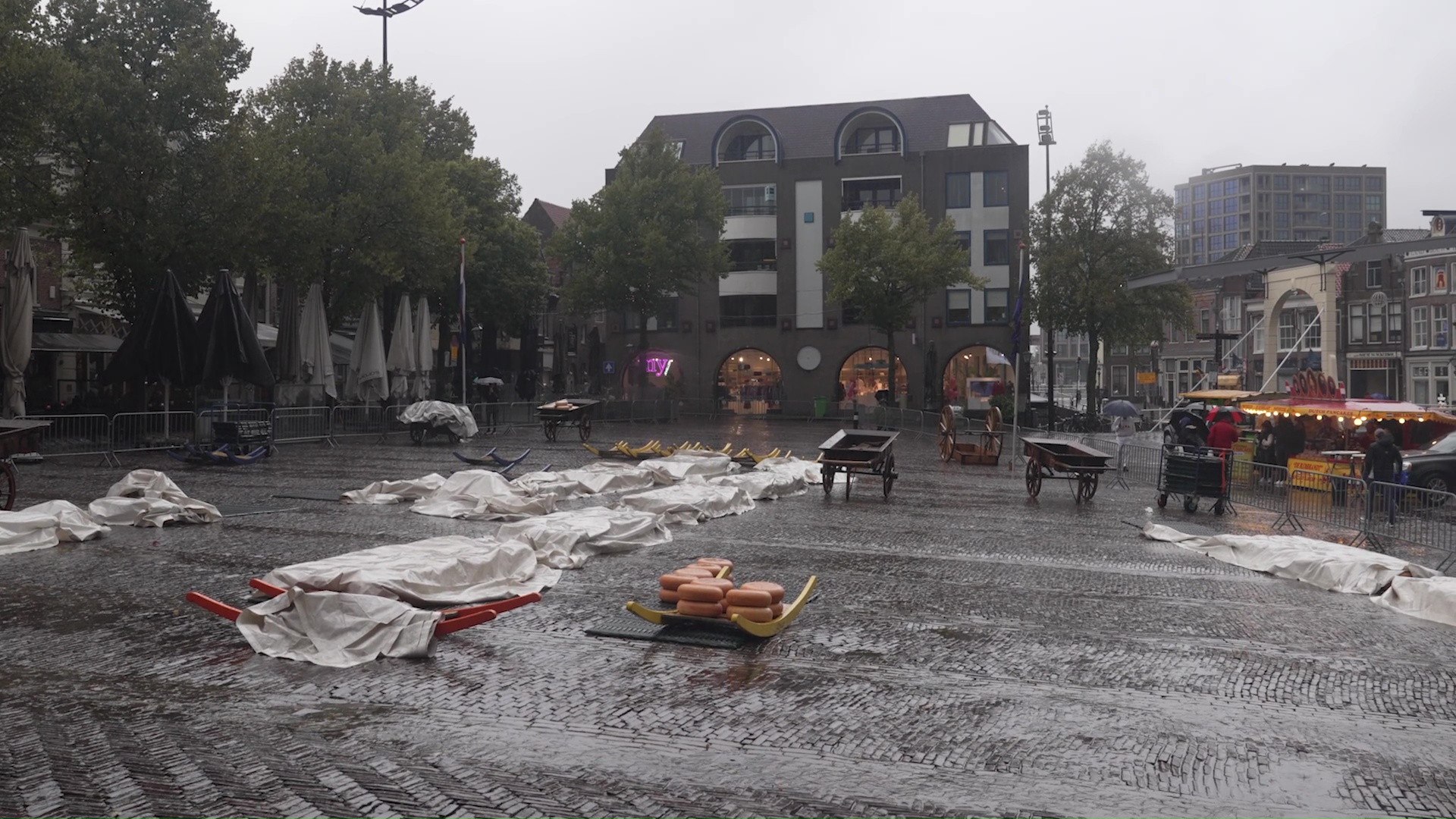 Nat marktplein met afgedekte kraampjes, natte straatstenen en een stapel kazen op een sleden in de regen.