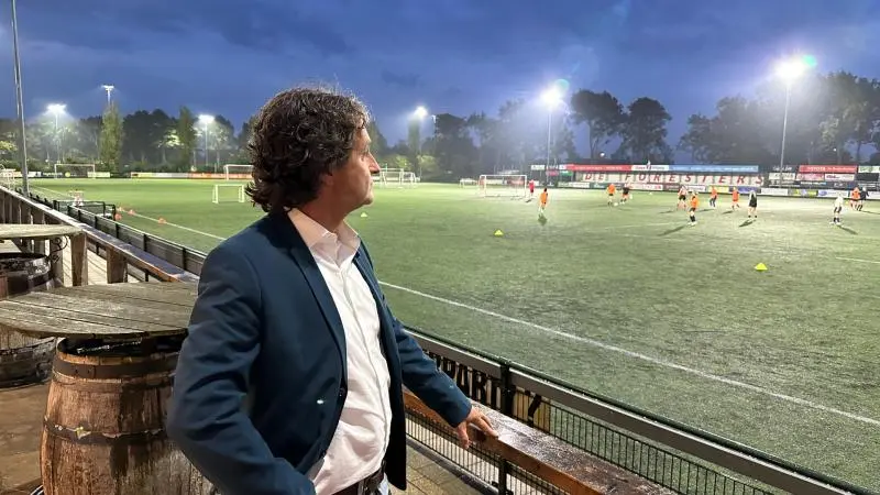 Man met blauwe blazer kijkt naar een voetbaltraining op een verlicht voetbalveld.