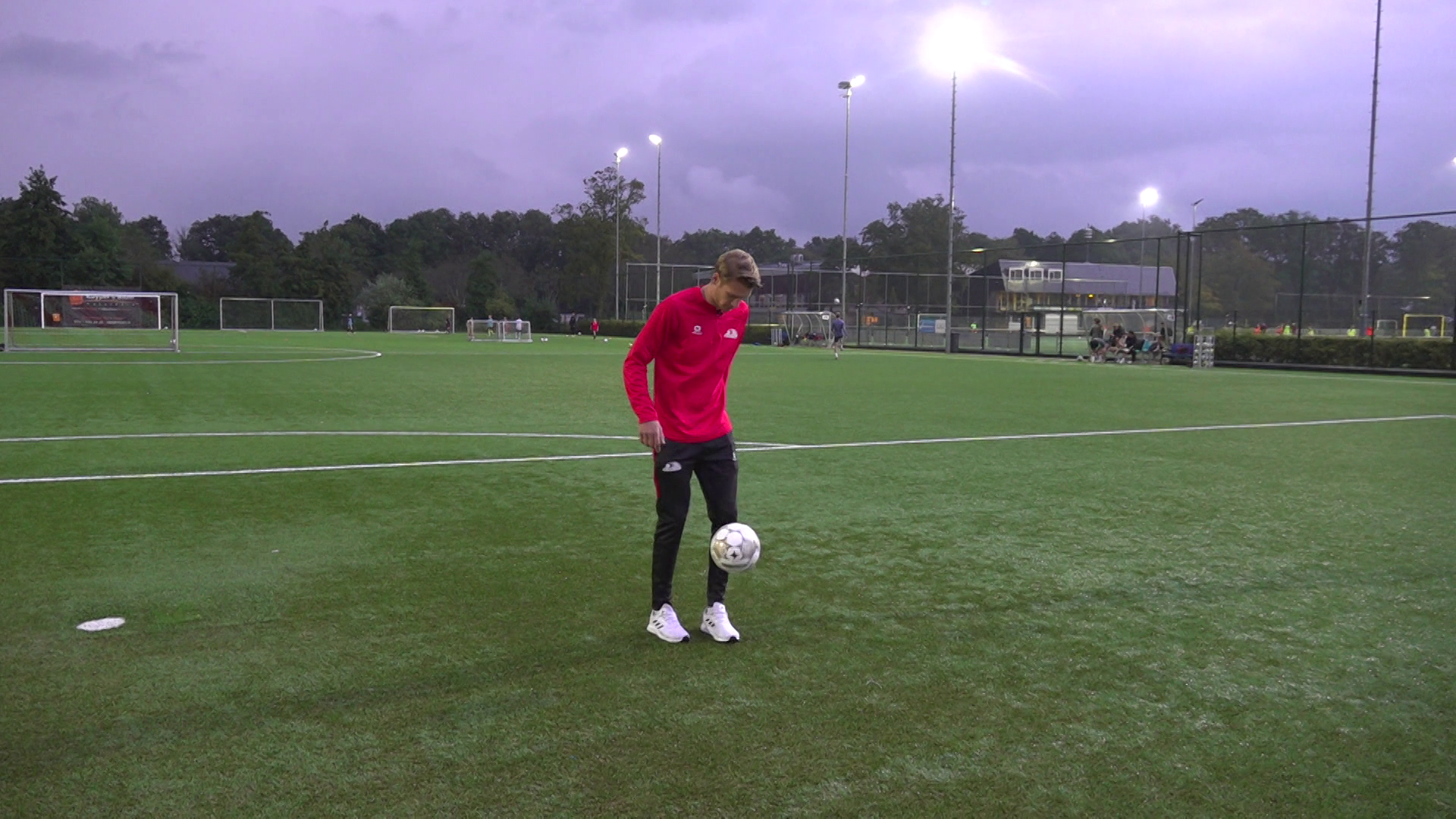 Voetballer in rood shirt en zwarte broek doet een truc met een voetbal op een verlicht kunstgrasveld.