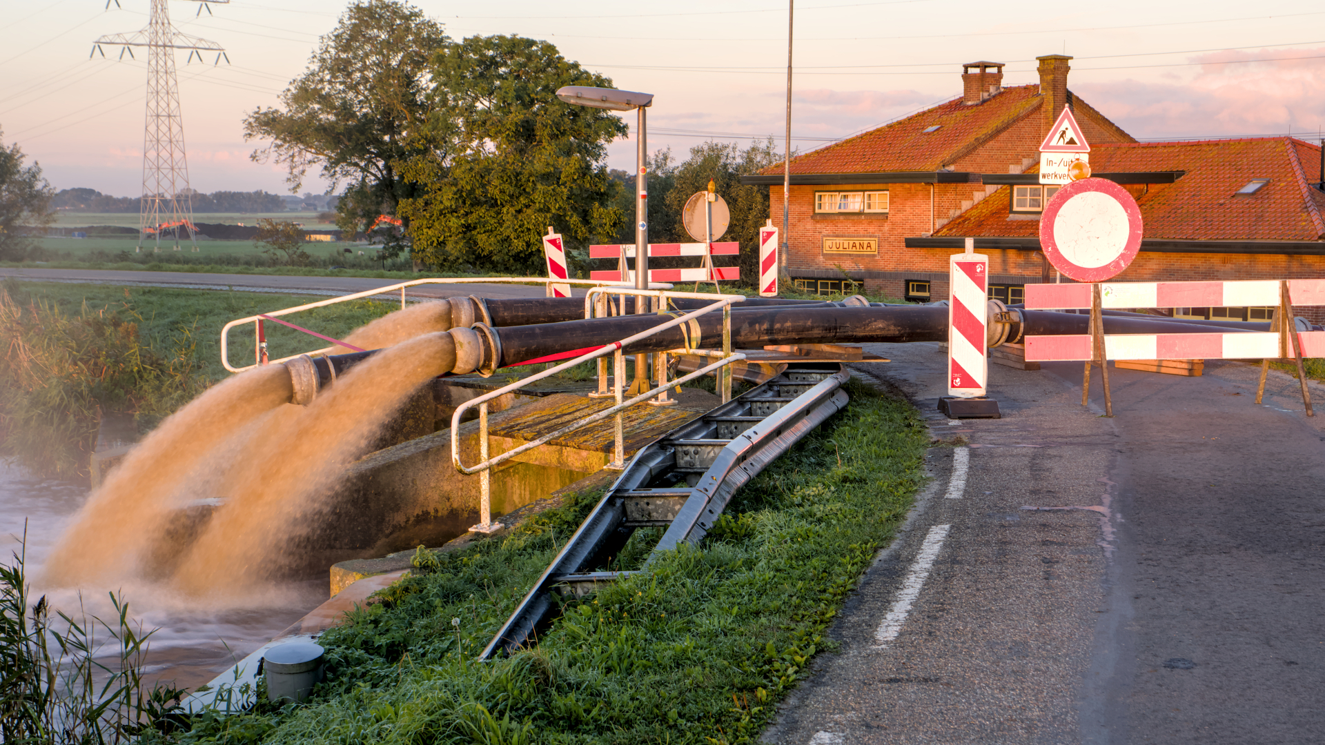 Water wordt door grote pompen afgevoerd langs een weg die is afgesloten met waarschuwingsborden en barrières. Op de achtergrond staat een gebouw met een rood dak en de naam "JULIANA".