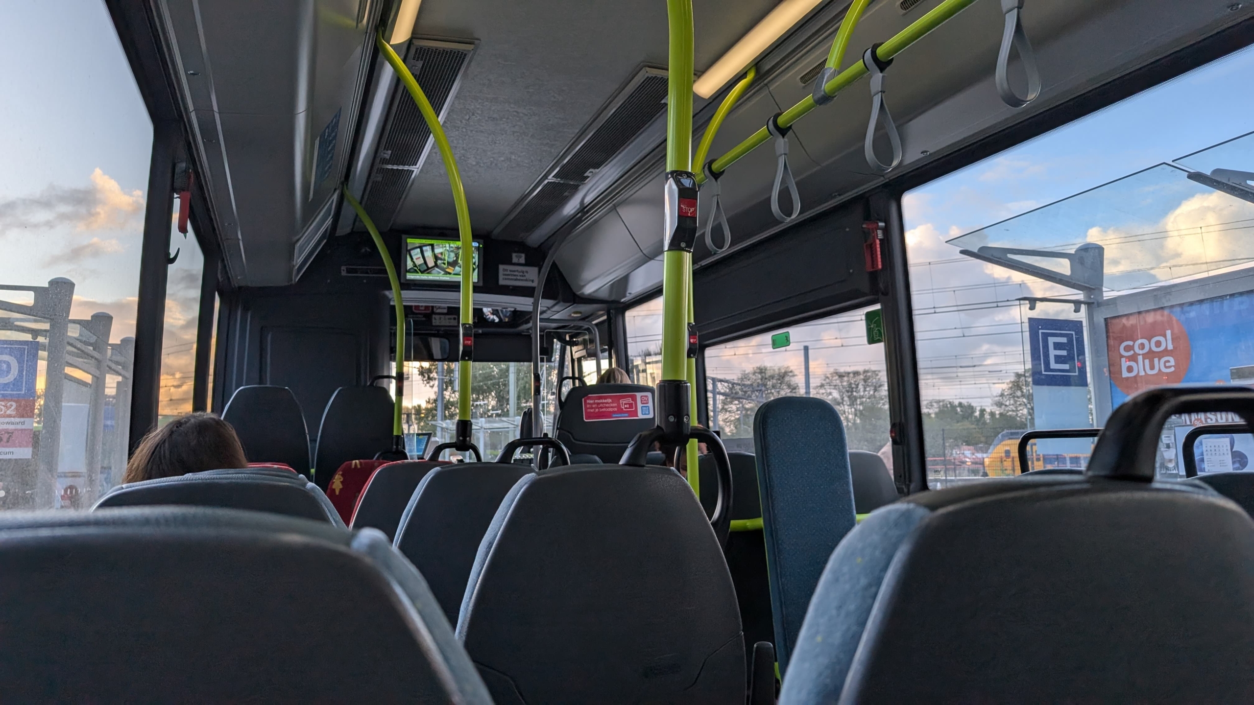 Binnenzicht van een lege bus met enkele passagiers en opvallende groene relingen. Door het raam zijn een parkeerbord en een Coolblue gebouw te zien.