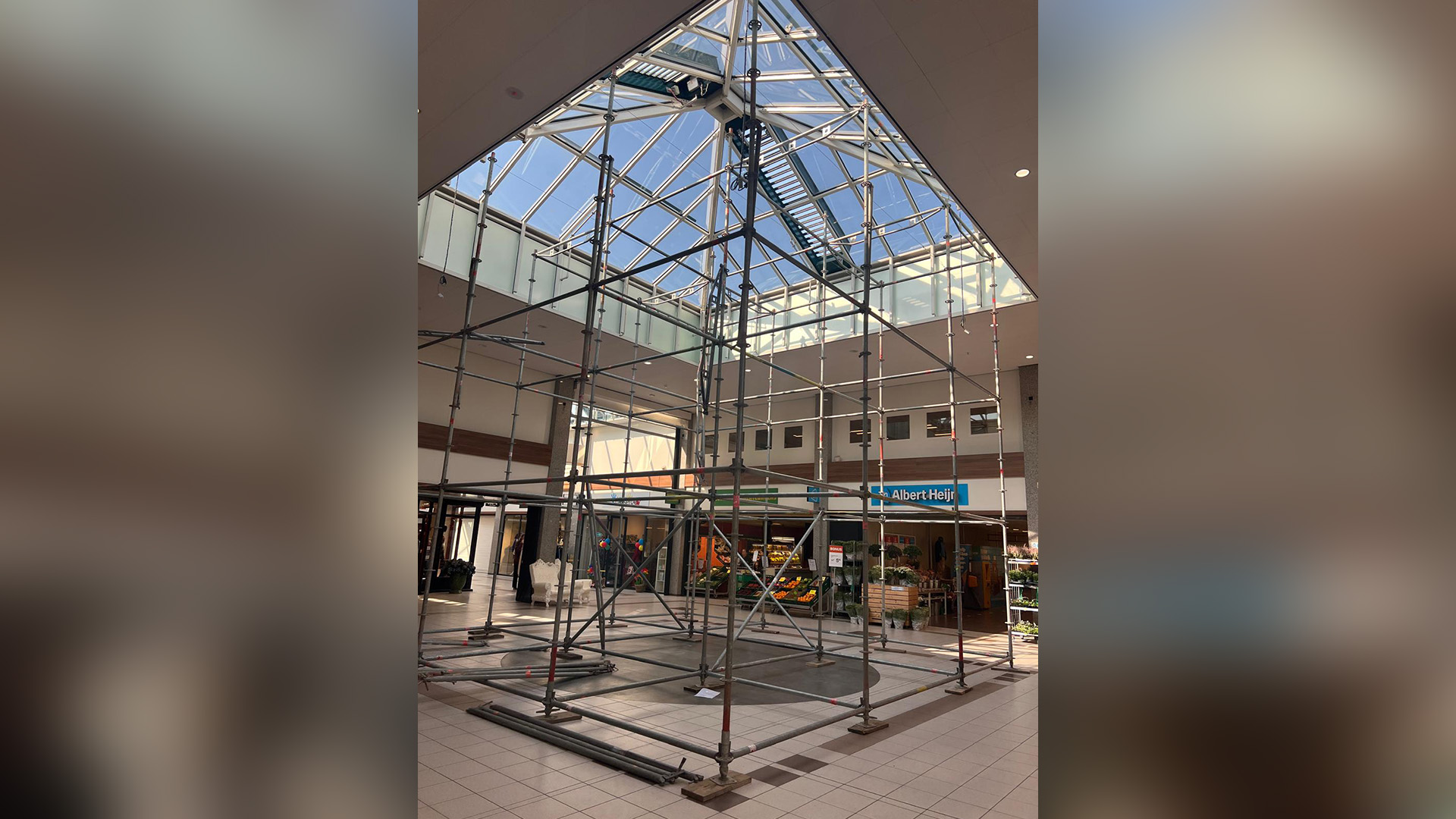 Binnenkant van een winkelcentrum met stalen steigers onder een glazen dakraam, op de achtergrond de ingang van een Albert Heijn supermarkt.