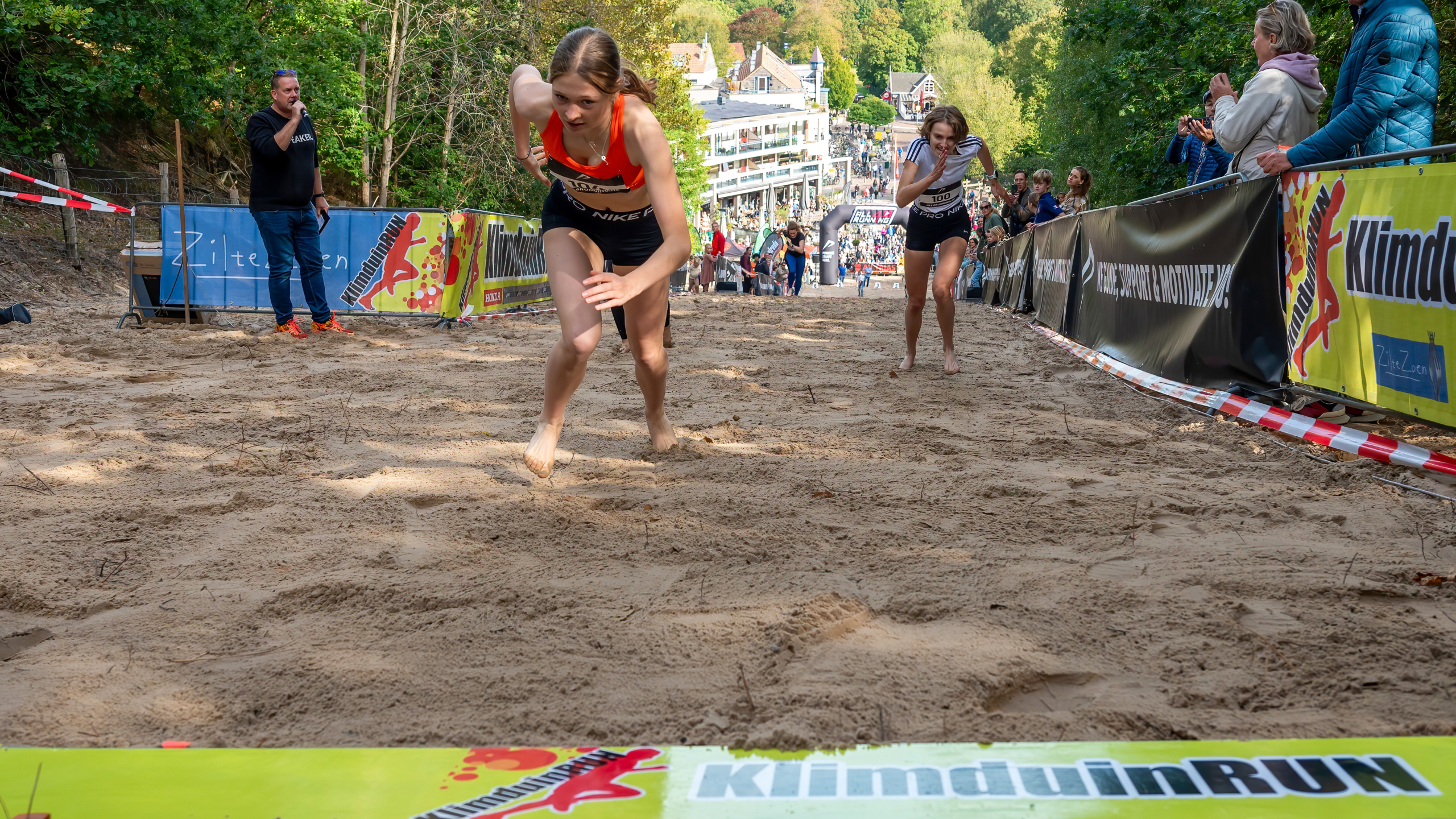 Twee vrouwen rennen op een zandheuvel tijdens de KlimduinRUN, met toeschouwers langs de zijlijn en een man met een microfoon.