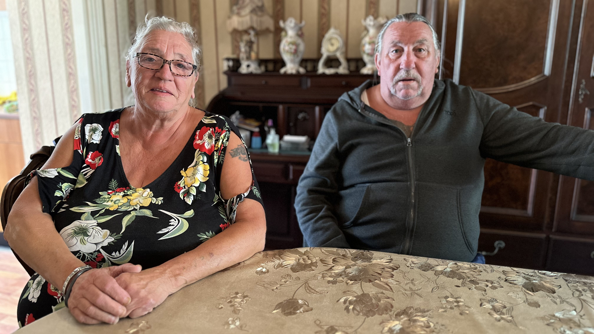 Twee oudere mensen zitten aan een tafel binnenshuis, de ene met een kleurrijke bloemenjurk en de andere met een donker vest.