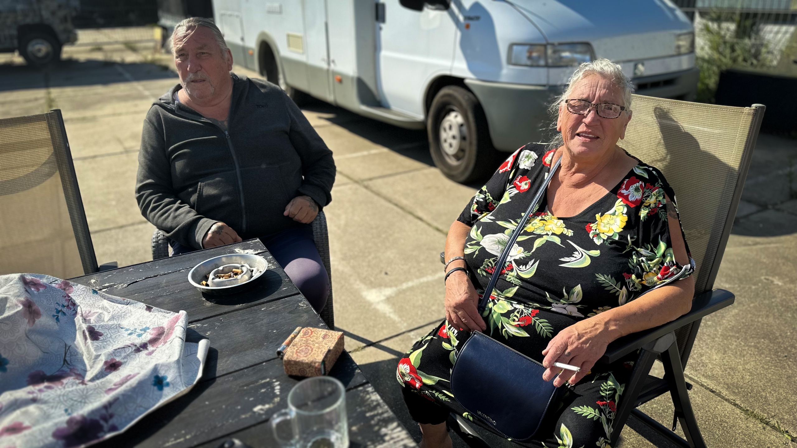 Twee oudere mensen zittend buiten bij een tafel; man in grijze trui, vrouw in bloemrijke jurk met een sigaret en een blauwe tas; op de achtergrond een camper.