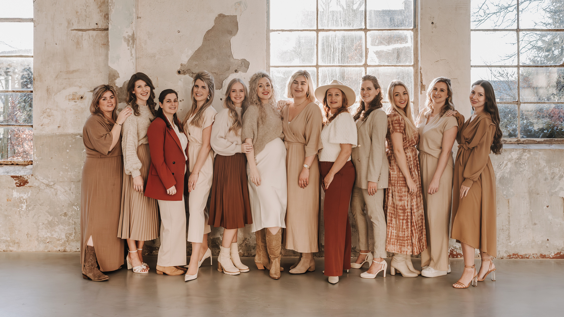 Groepsfoto van elf vrouwen in neutrale kleding staand in een industriële ruimte.