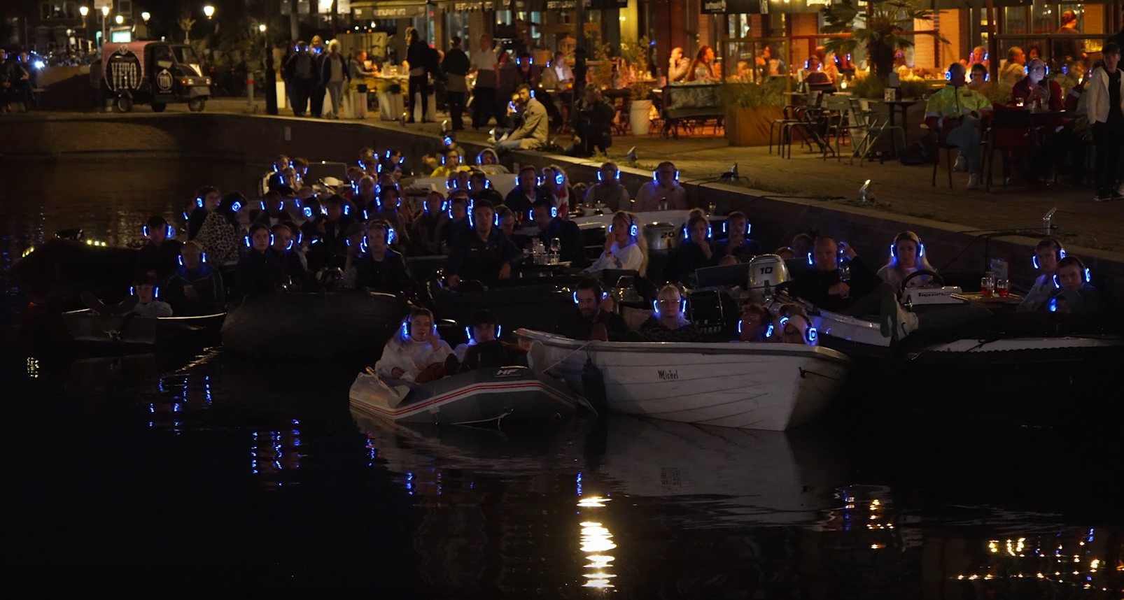 Mensen met oplichtende koptelefoons die 's avonds in boten op een gracht zitten, terwijl anderen op de kade toekijken.