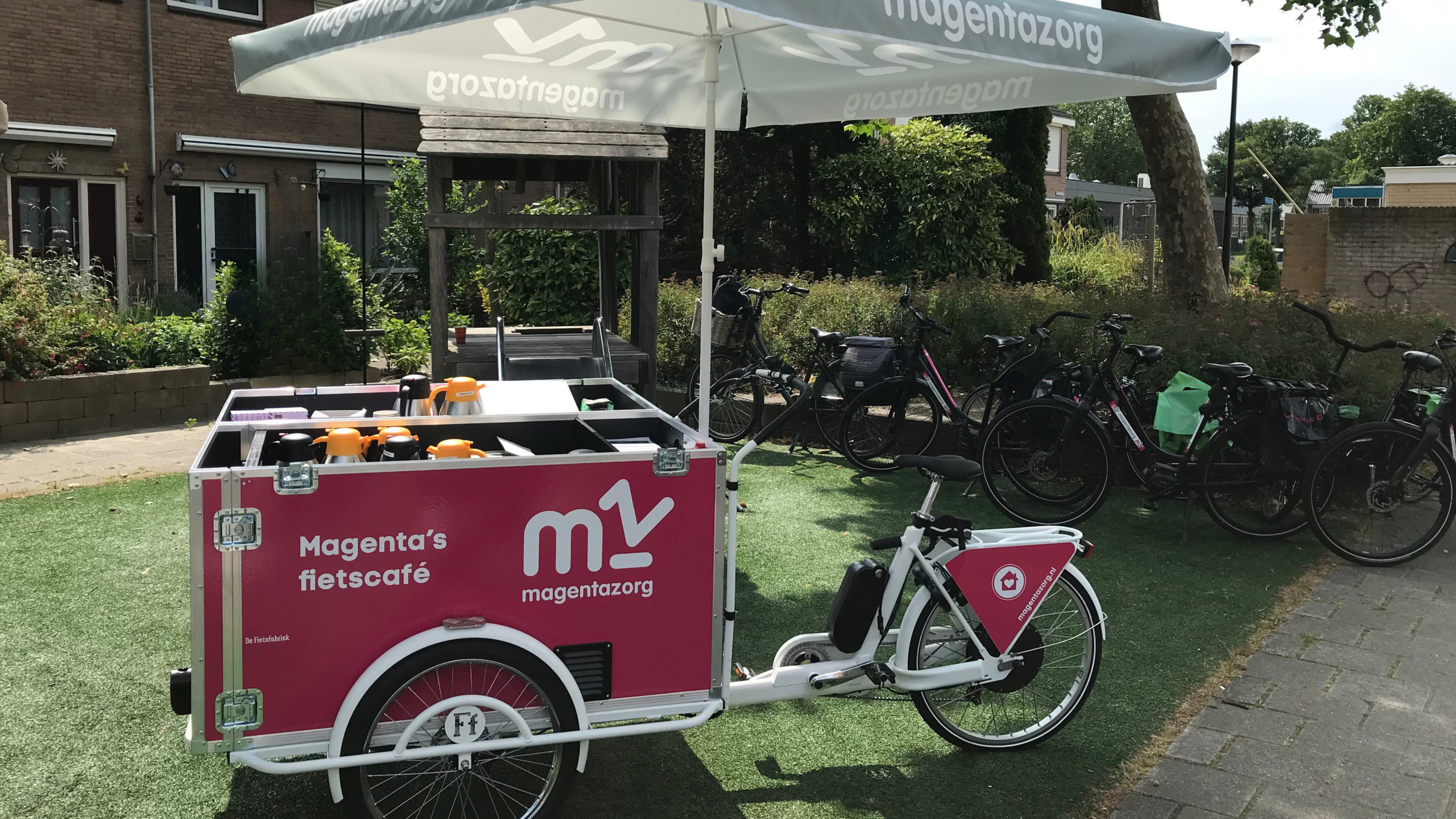 Bakfiets met opschrift "Magenta's fietscafé" onder een parasol, geparkeerd in een woonwijk. Meerdere fietsen staan op de achtergrond.