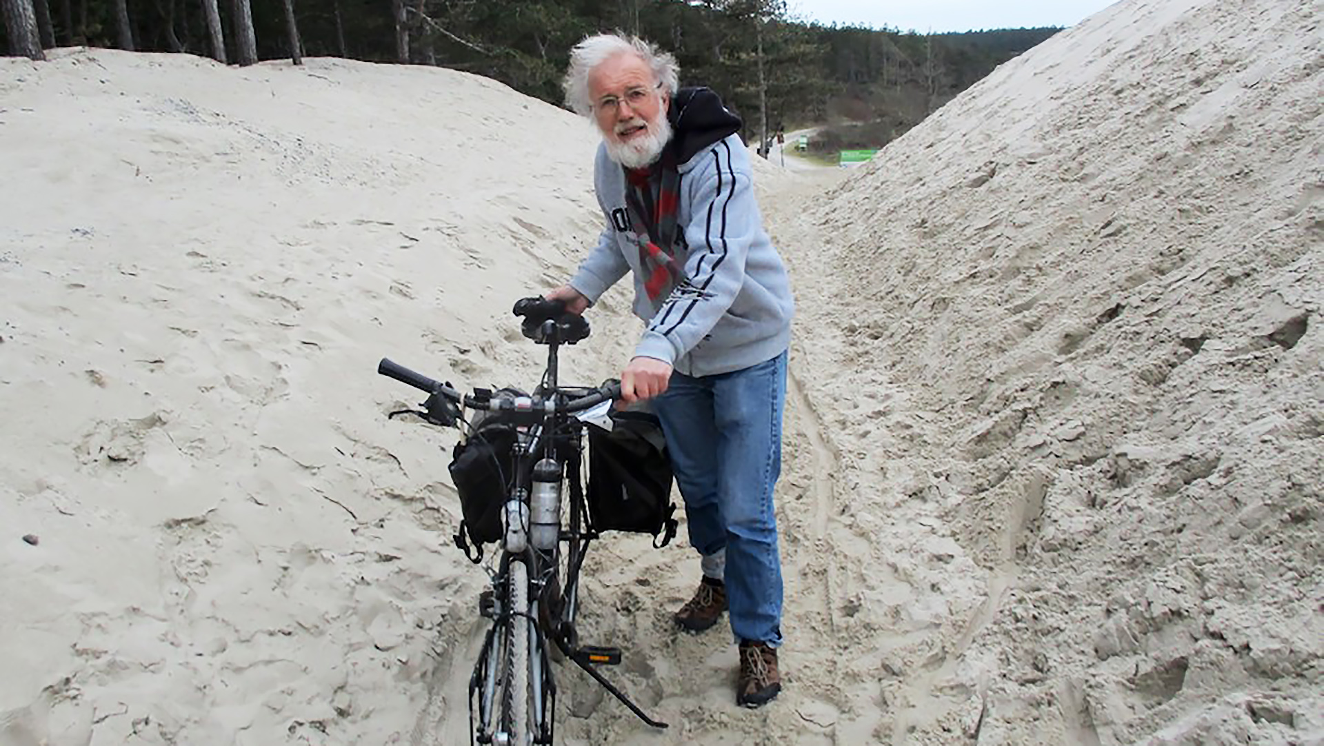 Man met grijs haar en een baard duwt een fiets door een zandduin.