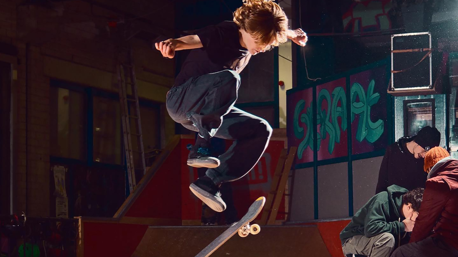 Jonge skateboarder maakt een stunt in een indoor skatepark met graffiti op de achtergrond en toeschouwers die toekijken.