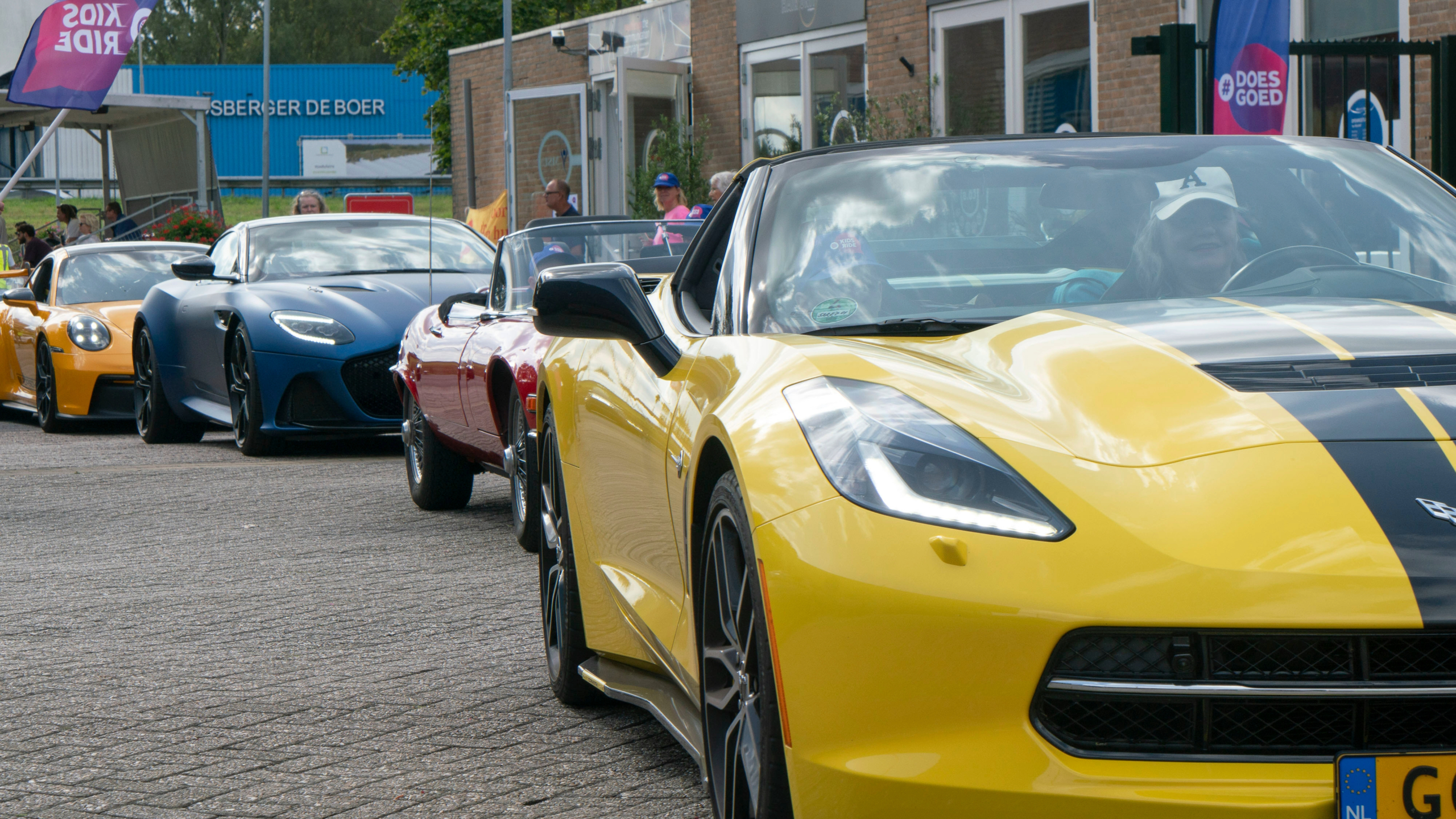 Rij van sportauto's, waaronder een gele Chevrolet Corvette, geparkeerd buiten een gebouw, mensen op de achtergrond.