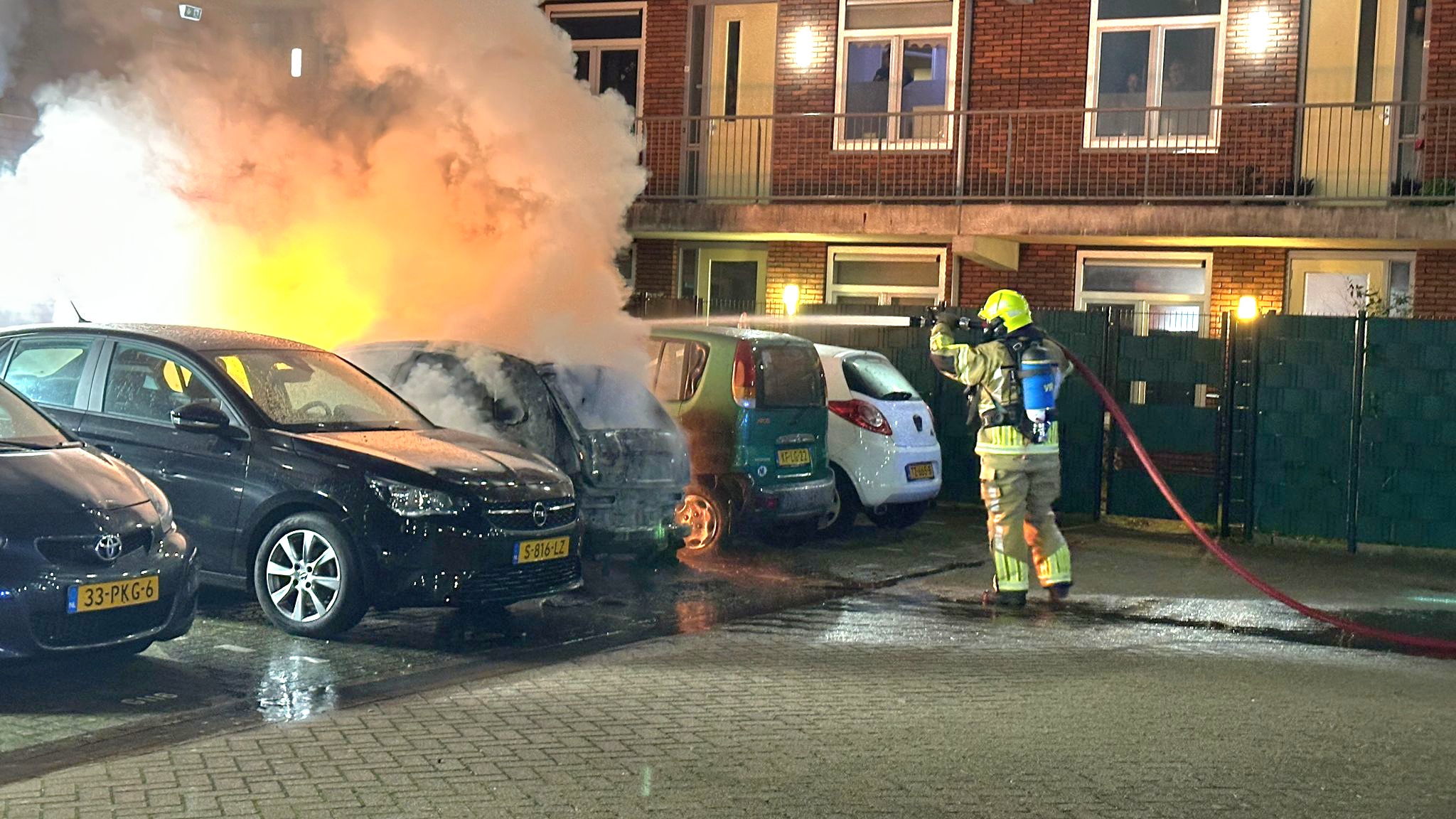 Brandweerman blust brandende auto op parkeerplaats naast andere geparkeerde voertuigen, dikke rookontwikkeling zichtbaar.