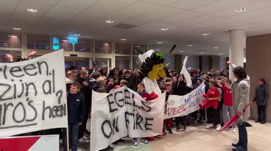 Een grote groep mensen met zelfgemaakte borden, waaronder een mascotte in een egelpak, staat bijeen in een lichte gang binnen een gebouw.