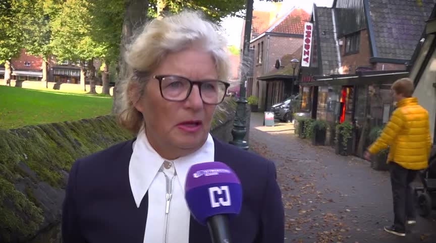 Vrouw met bril wordt geïnterviewd met microfoon in straat met bomen en winkels.