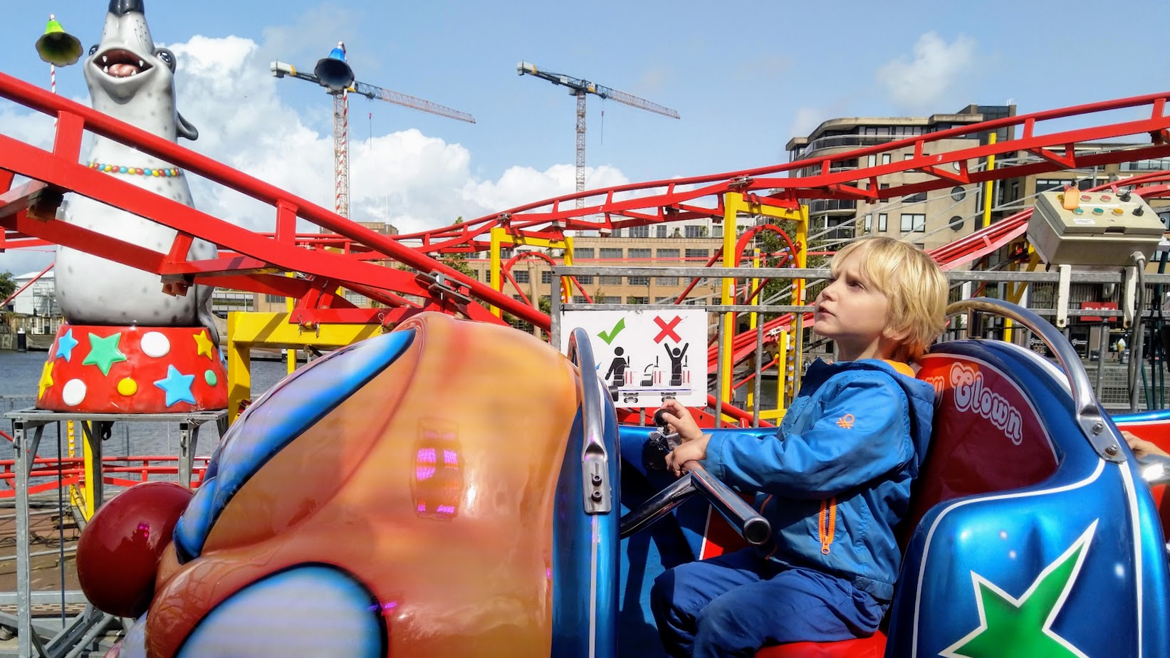 Een kind in een blauwe jas zit in een achtbaanwagen naast een kleurrijke clownzeehondfiguur.