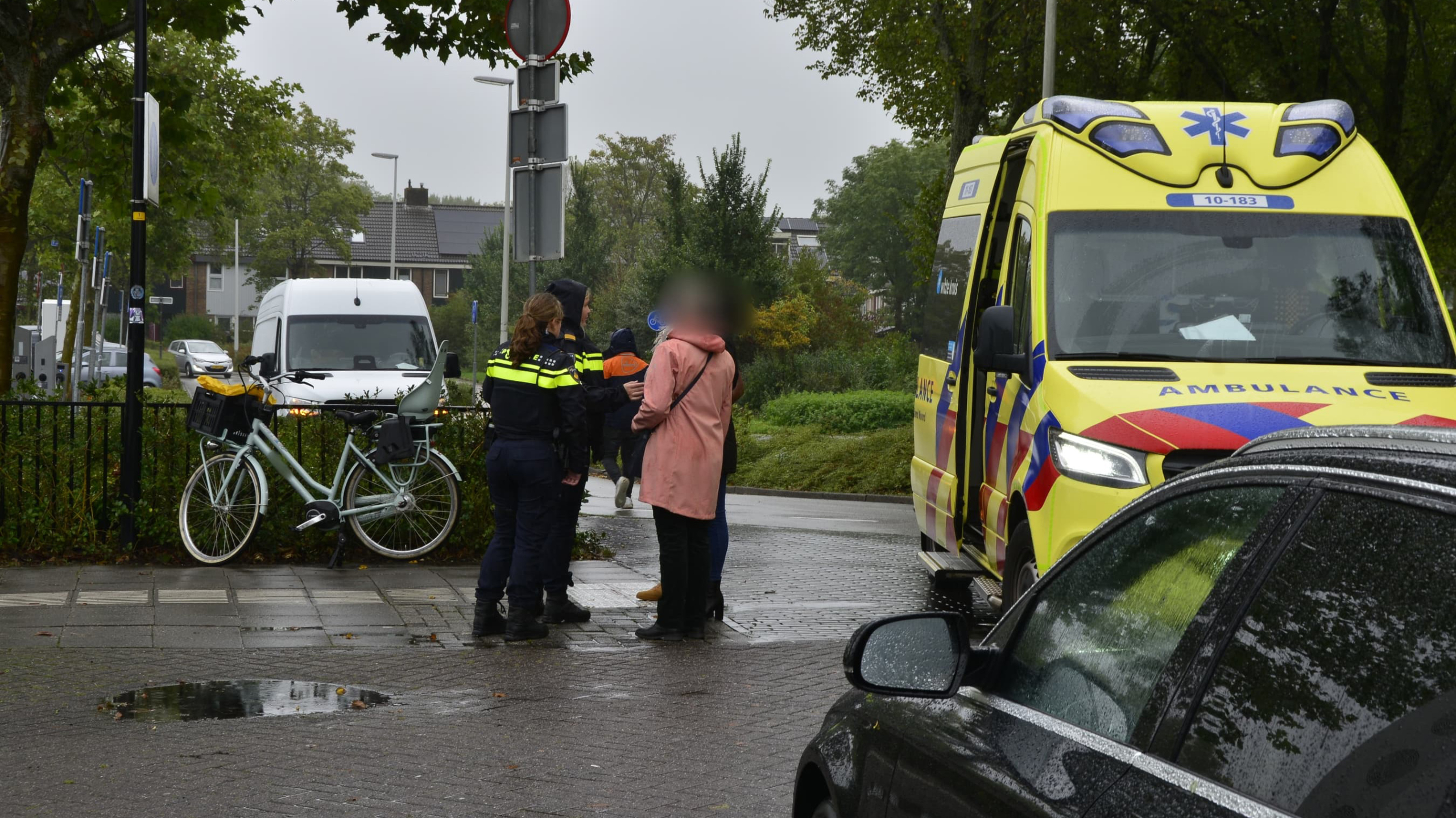 Politieagenten en mensen naast een ambulance op een regenachtige straat bij een fiets.