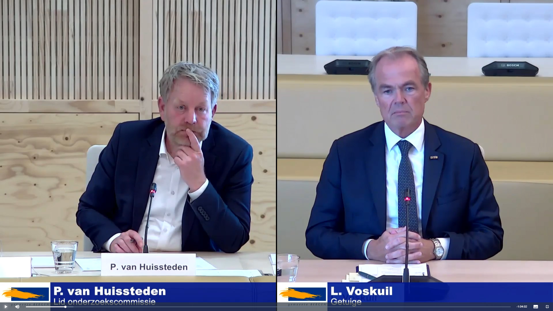 Twee mannen in pakken zitten aan een tafel met microfoons tijdens een vergadering. De namen "P. van Huisssteden" en "L. Voskuil" zijn te zien op naambordjes.