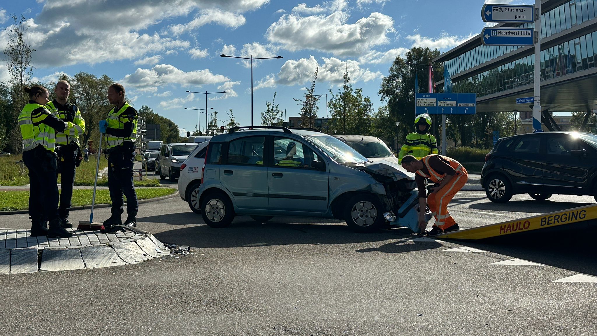 Een blauwe auto met schade aan de voorkant staat op een rotonde, omringd door politieagenten en bergingspersoneel.