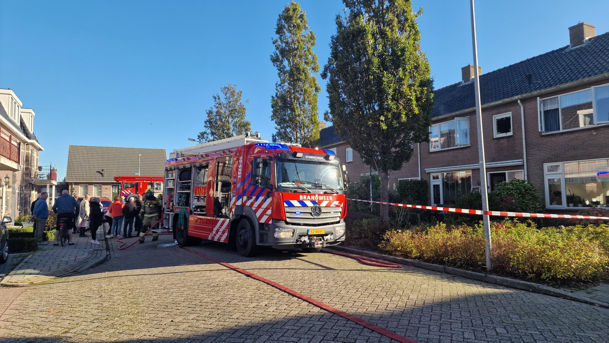 Brandweerwagen geparkeerd in een woonstraat, omringd door toeschouwers en politieafzetting, met een huis op de achtergrond.