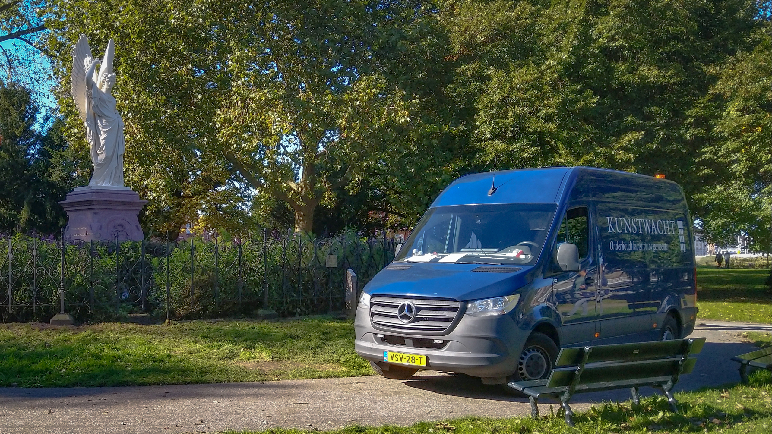 Een blauw busje van Kunstwacht naast een standbeeld van een engel in een park.