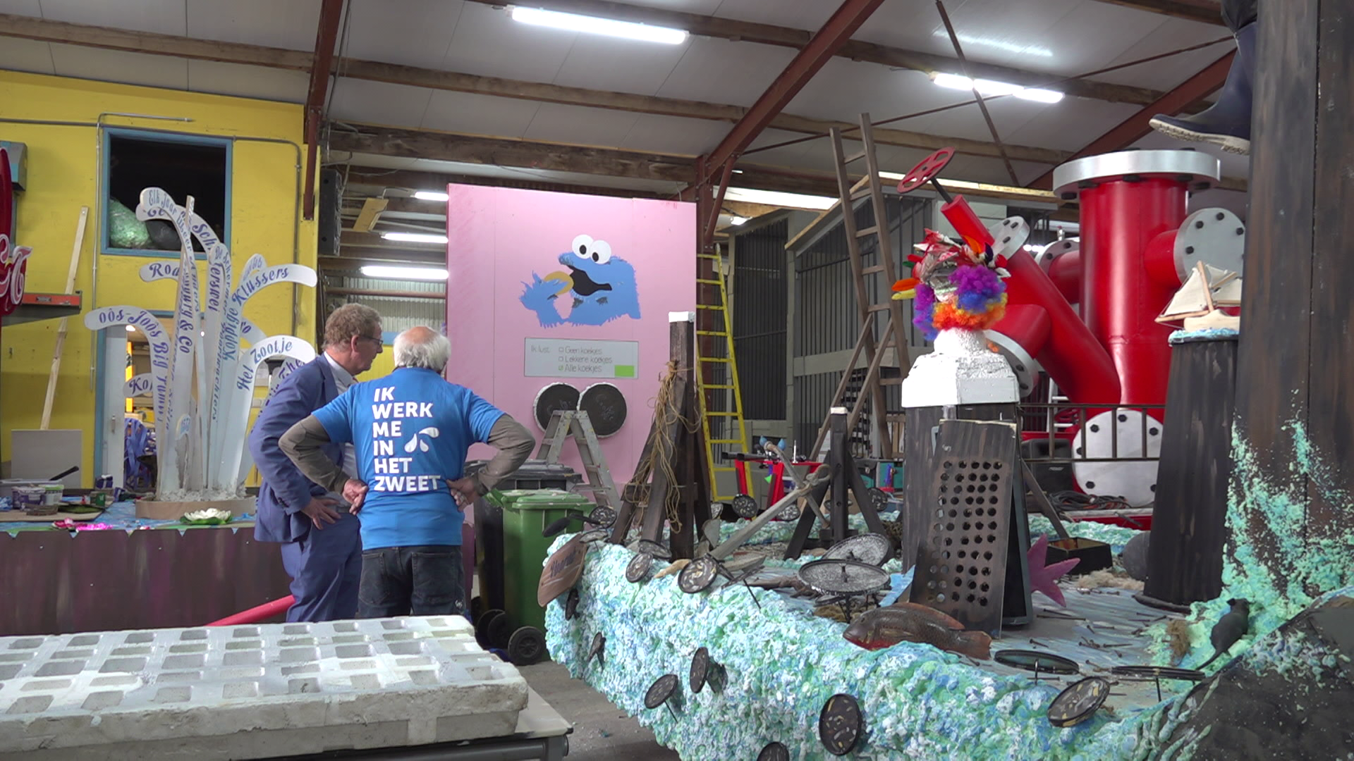 Een werkplaats met mensen die aan een kleurrijke carnavalswagen bouwen; op de achtergrond een roze paneel met een blauwe figuur en tekst op T-shirt van een persoon "IK WERK ME IN HET ZWEET".