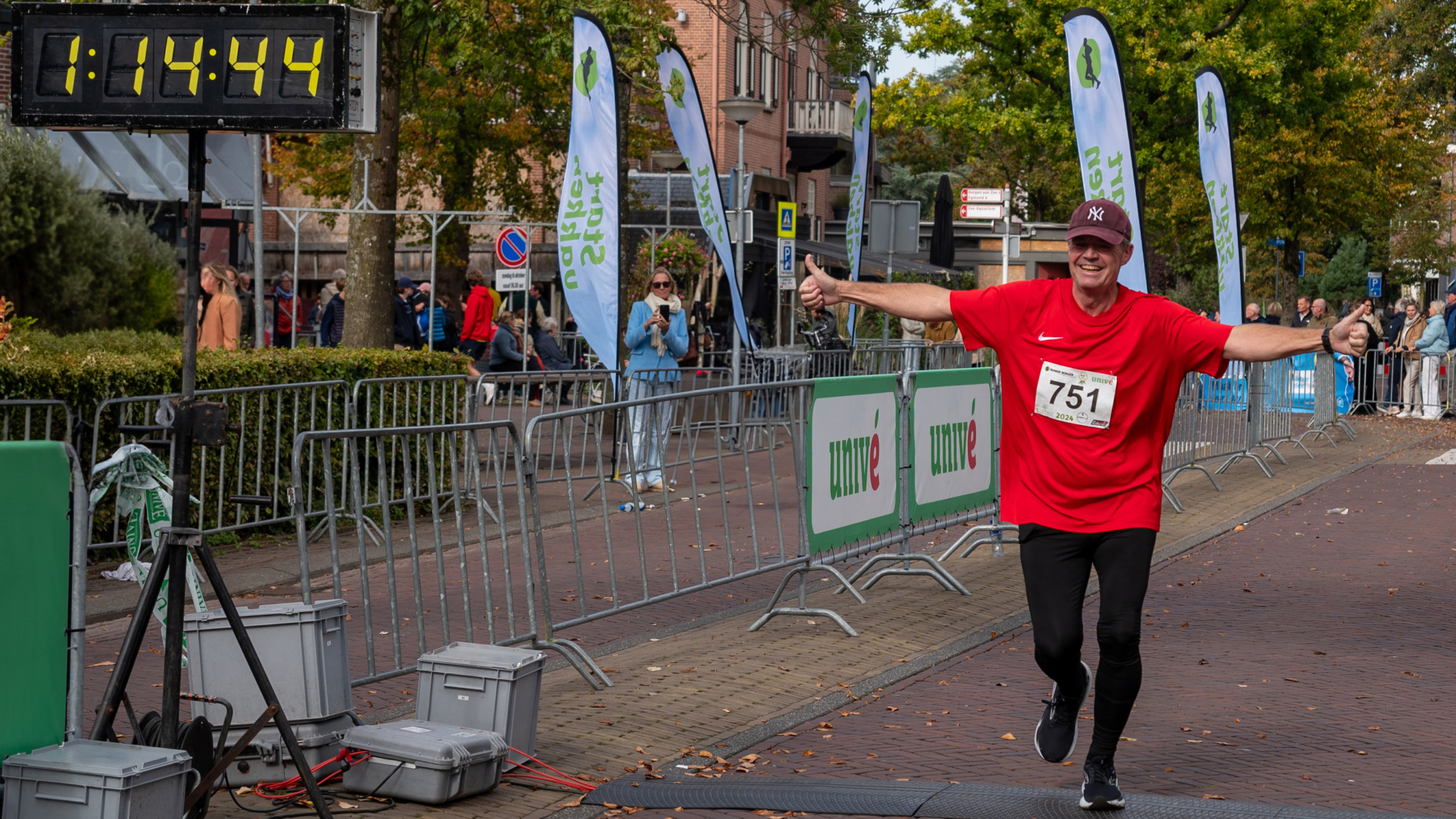 Hardloper in rood shirt met nummer 751 passeert de finishlijn tijdens een evenement, omringd door publiek en vlaggen. Een digitale timer toont 1:14:44.