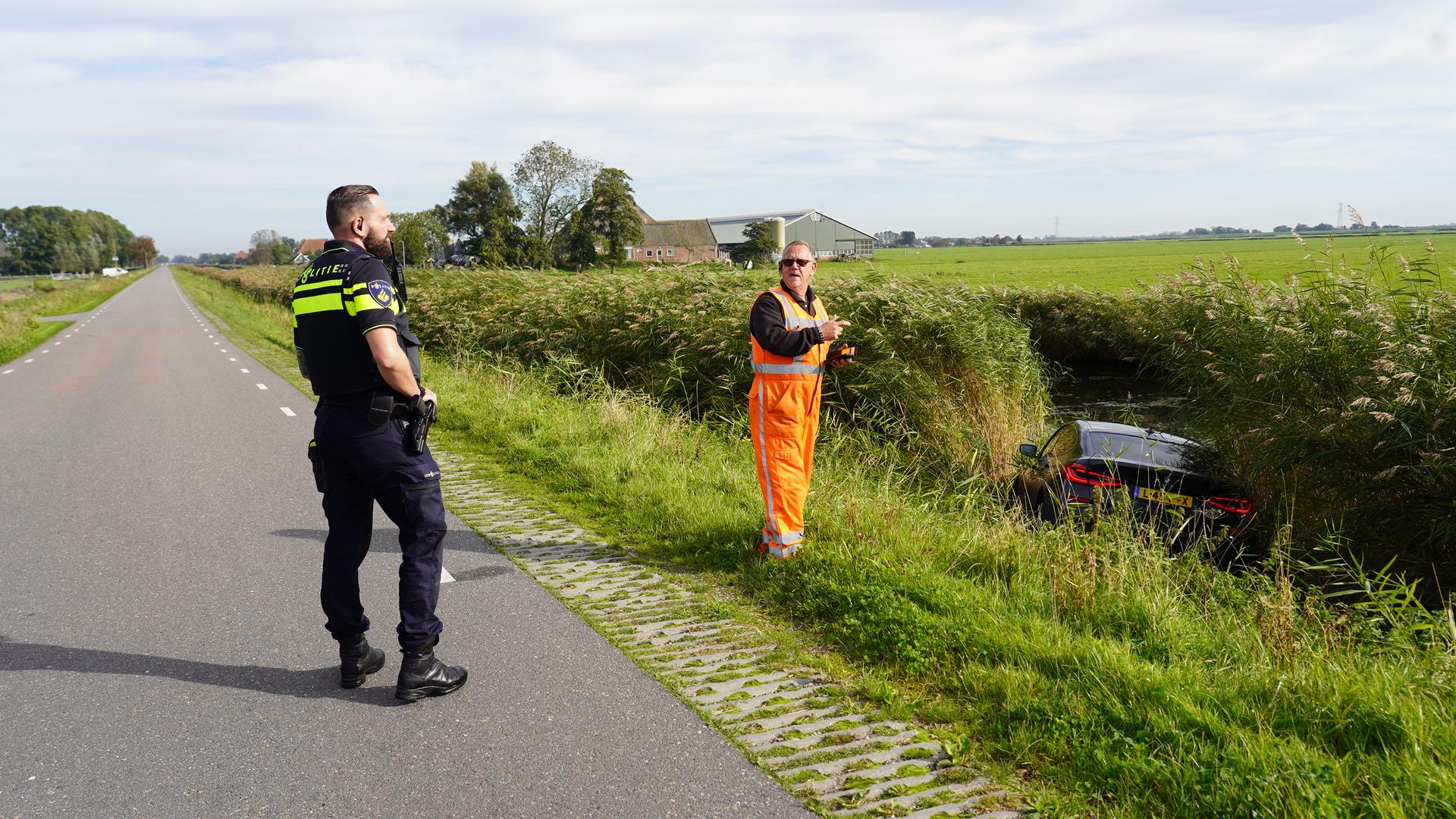 Politieagent en werker in oranje veiligheidskleding bij een auto die in een sloot langs de weg is beland, met velden en een boerderij op de achtergrond.