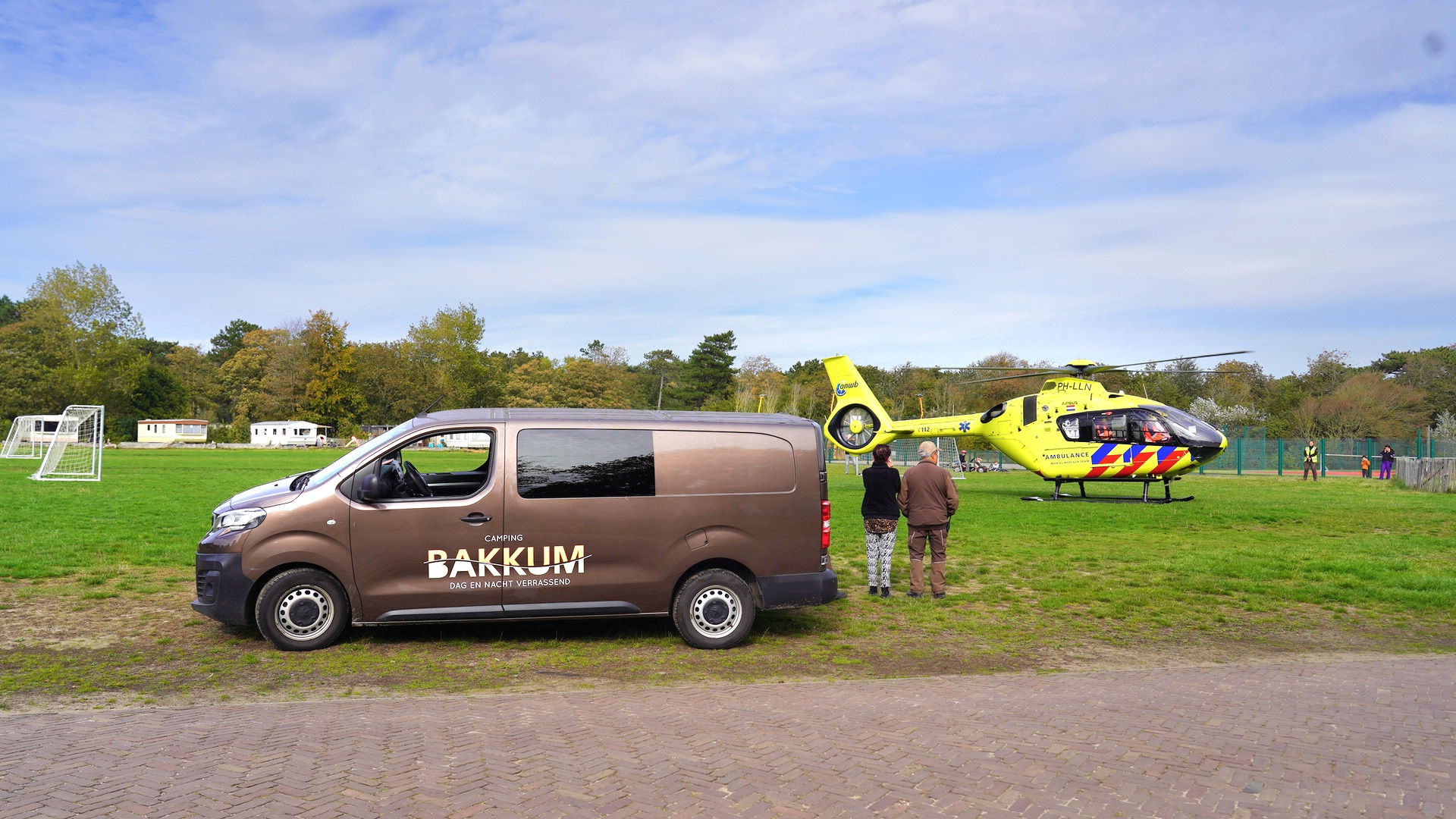 Bruine bestelwagen met opschrift "Camping Bakkum" geparkeerd naast een gele traumahelikopter op een grasveld, met mensen op de achtergrond.