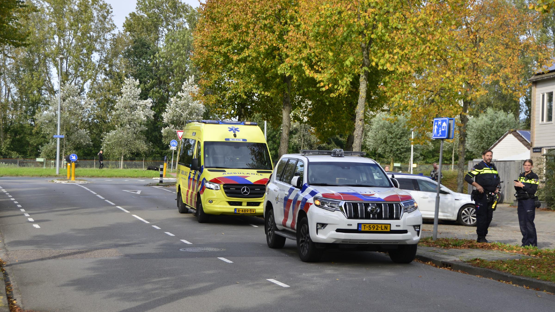 Twee politieagenten naast politieauto en ambulance op een straat met bomen en een huis.