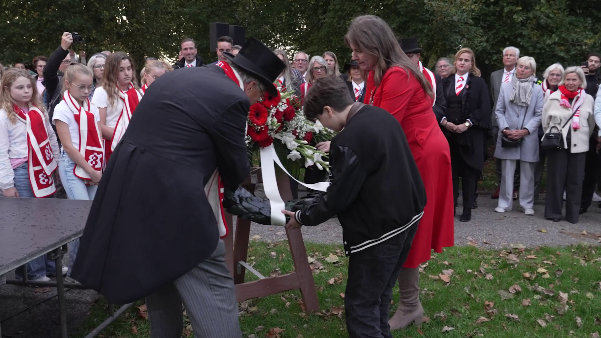 Een man en een jongen leggen een bloemenkrans neer tijdens een ceremonie, omringd door mensen.