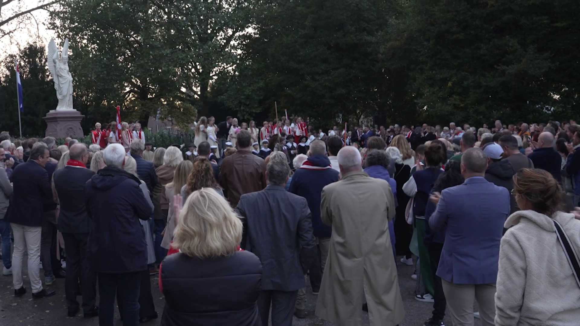 Een menigte verzameld rondom een standbeeld, met mensen in rode kledij op de achtergrond.