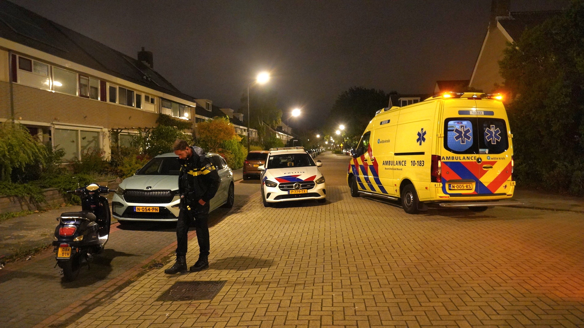 Politie- en ambulancedienstvoertuigen 's nachts op straat, met een politieman die naast een geparkeerde scooter en auto staat.