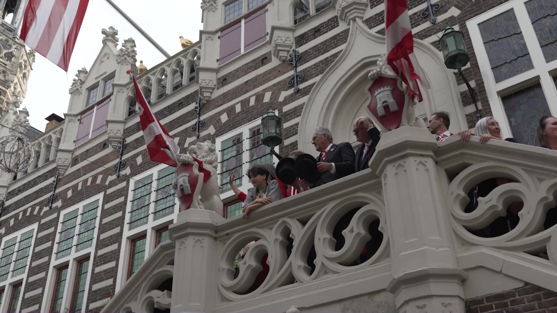 Groep mensen op een balkon van een historisch gebouw met rood-witte vlaggen.