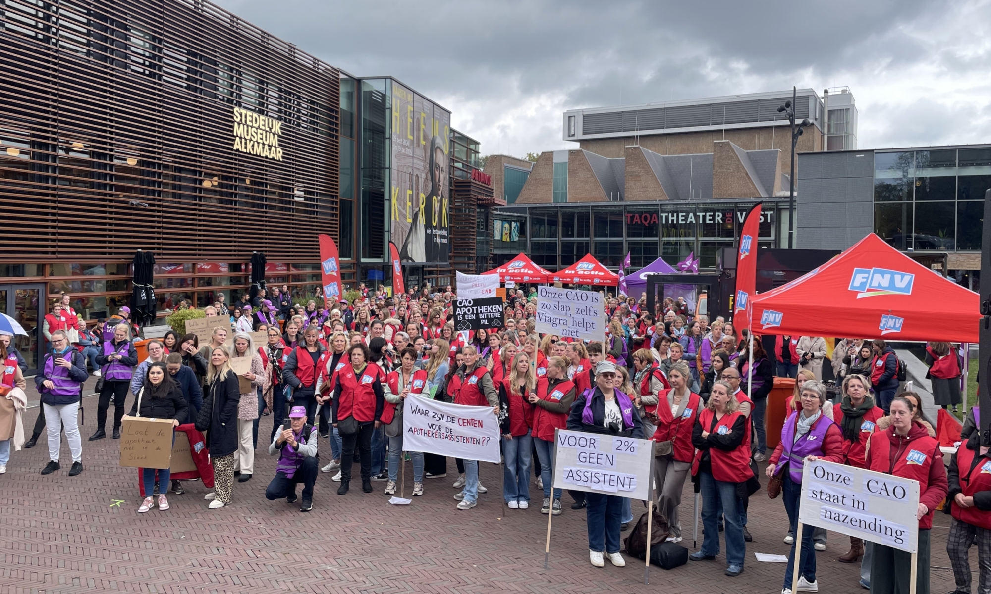 Een grote groep mensen verzameld voor een demonstratie bij het Stedelijk Museum Alkmaar, met FNV-tenten en spandoeken over arbeidsvoorwaarden.