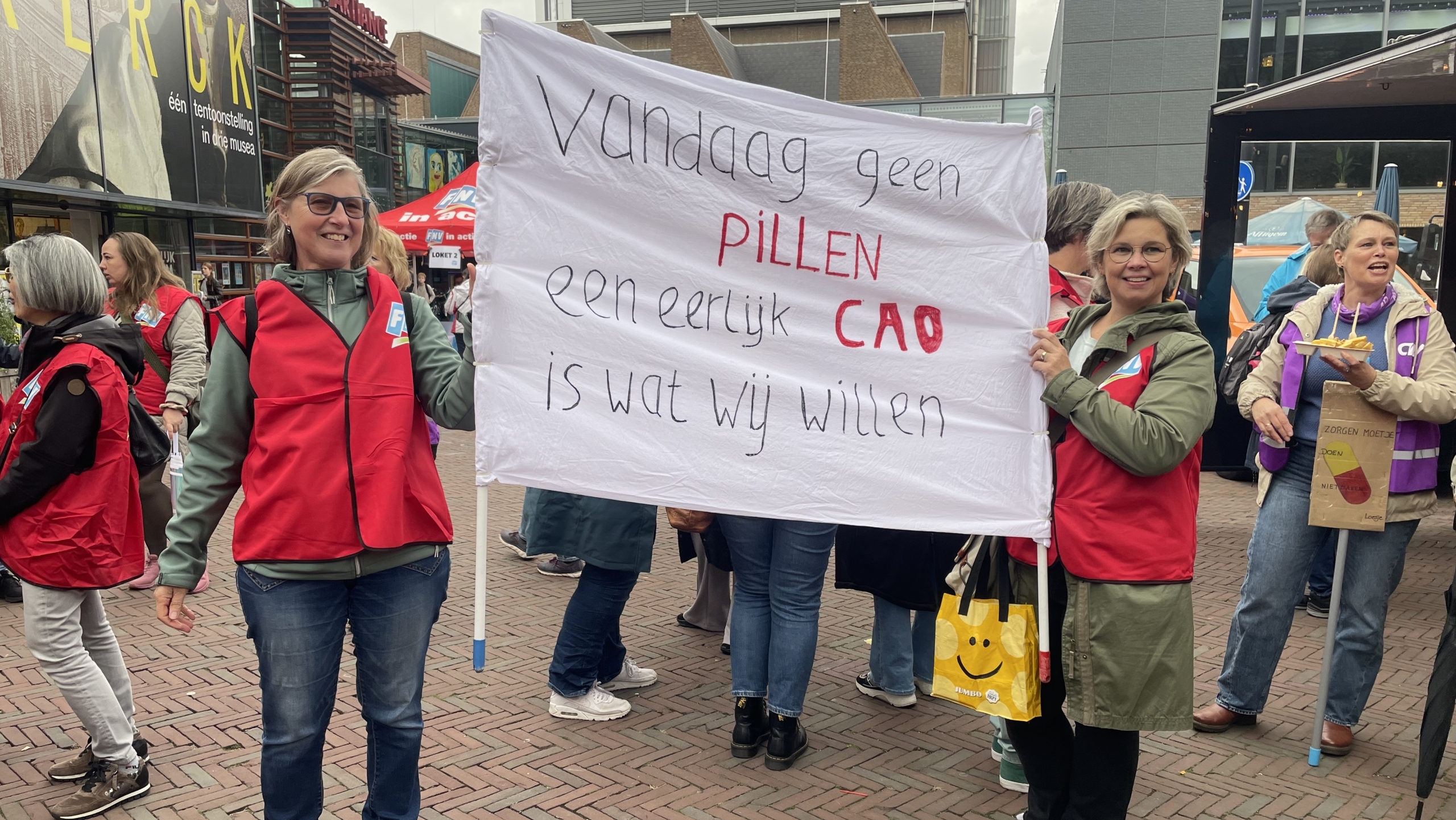 Twee vrouwen in rode hesjes houden een spandoek vast met de tekst "Vandaag geen pillen, een eerlijk CAO is wat wij willen" tijdens een protest.