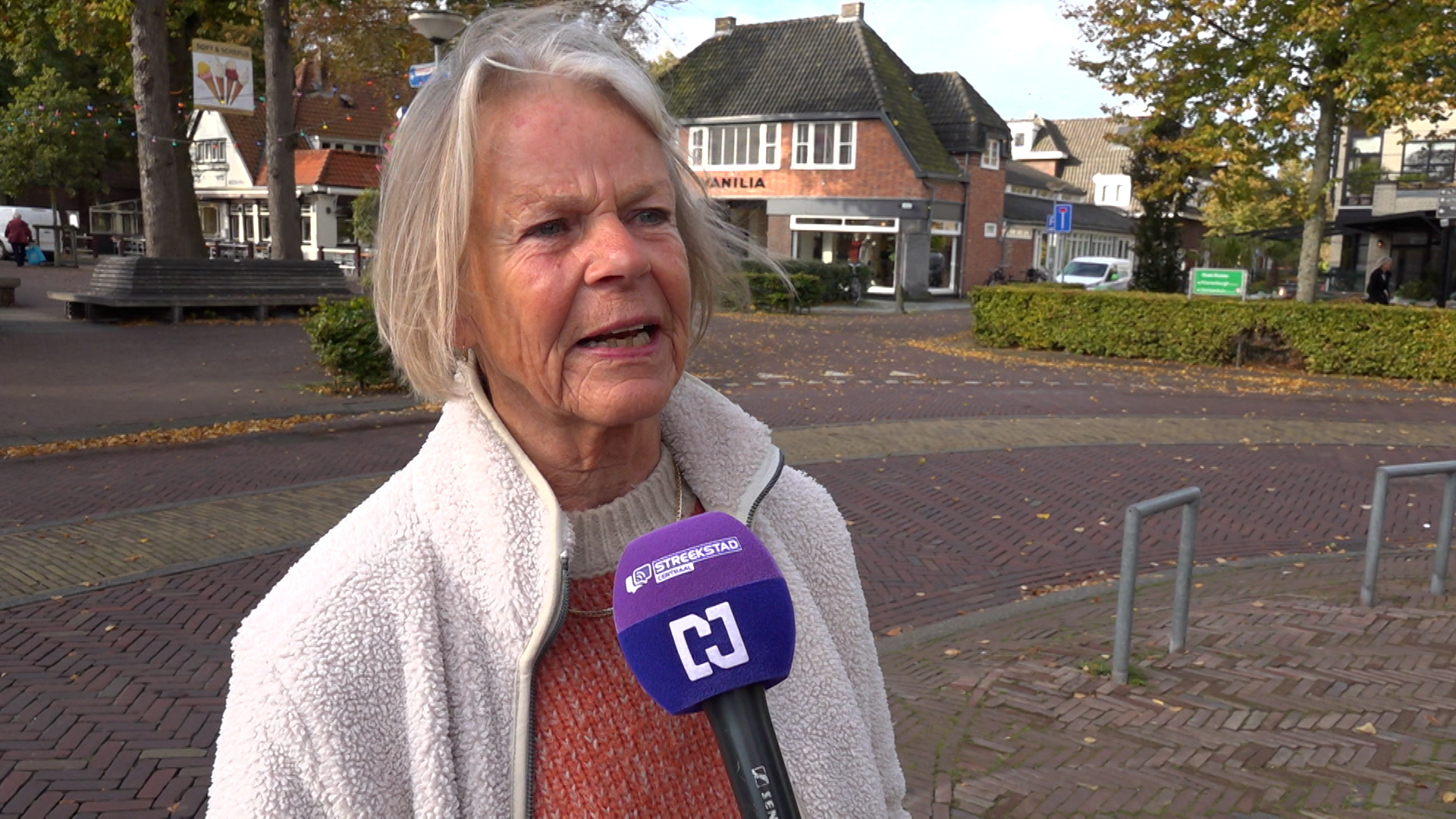 Oudere vrouw met kort grijs haar wordt geïnterviewd door een persmicrofoon in een stadsplein met bakstenen gebouwen en herfstbomen op de achtergrond.