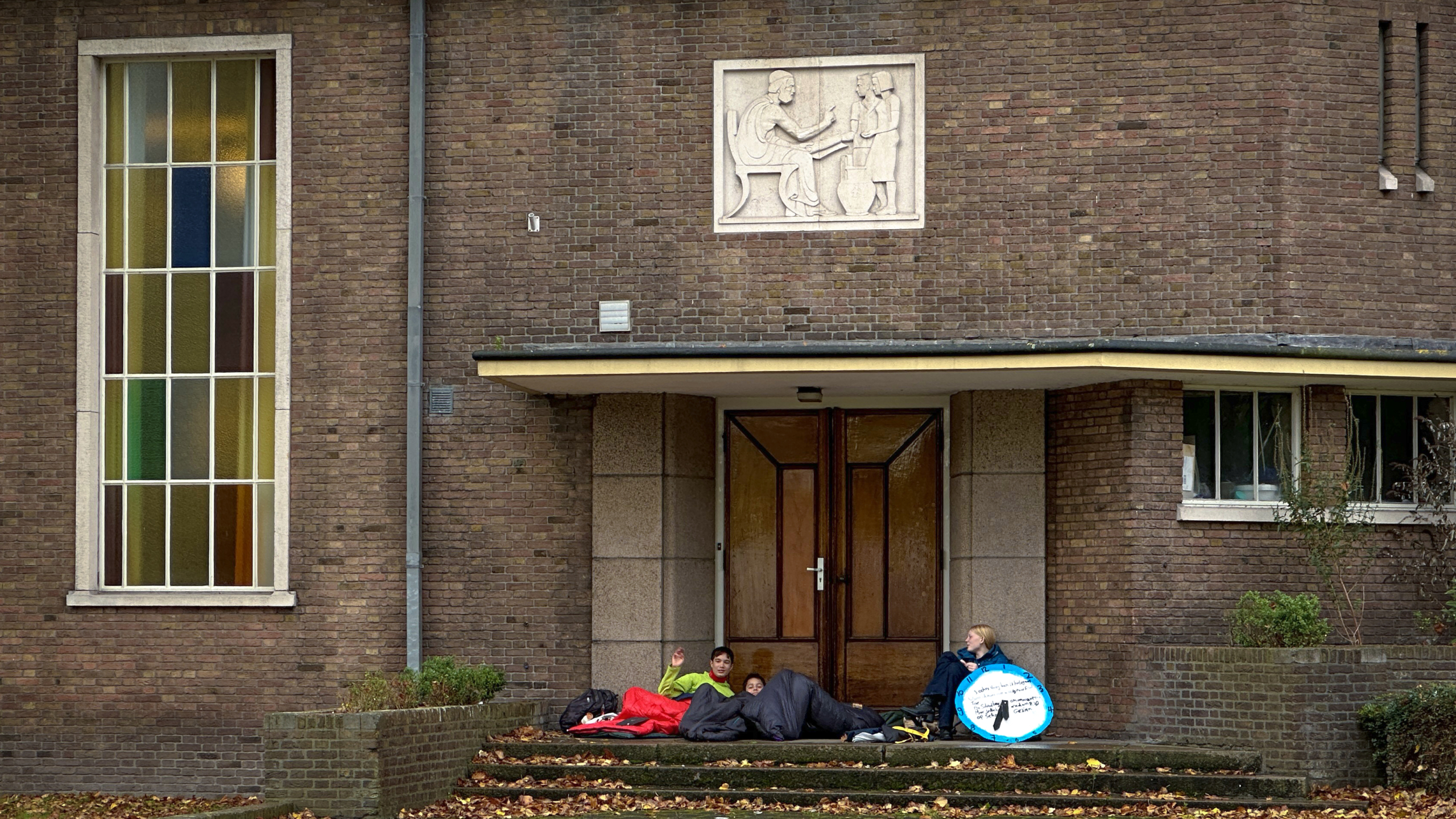 Twee mensen in slaapzakken en een persoon met een bord zitten op de stoep voor een gebouw met glazen ramen en een stenen reliëf boven de deur.