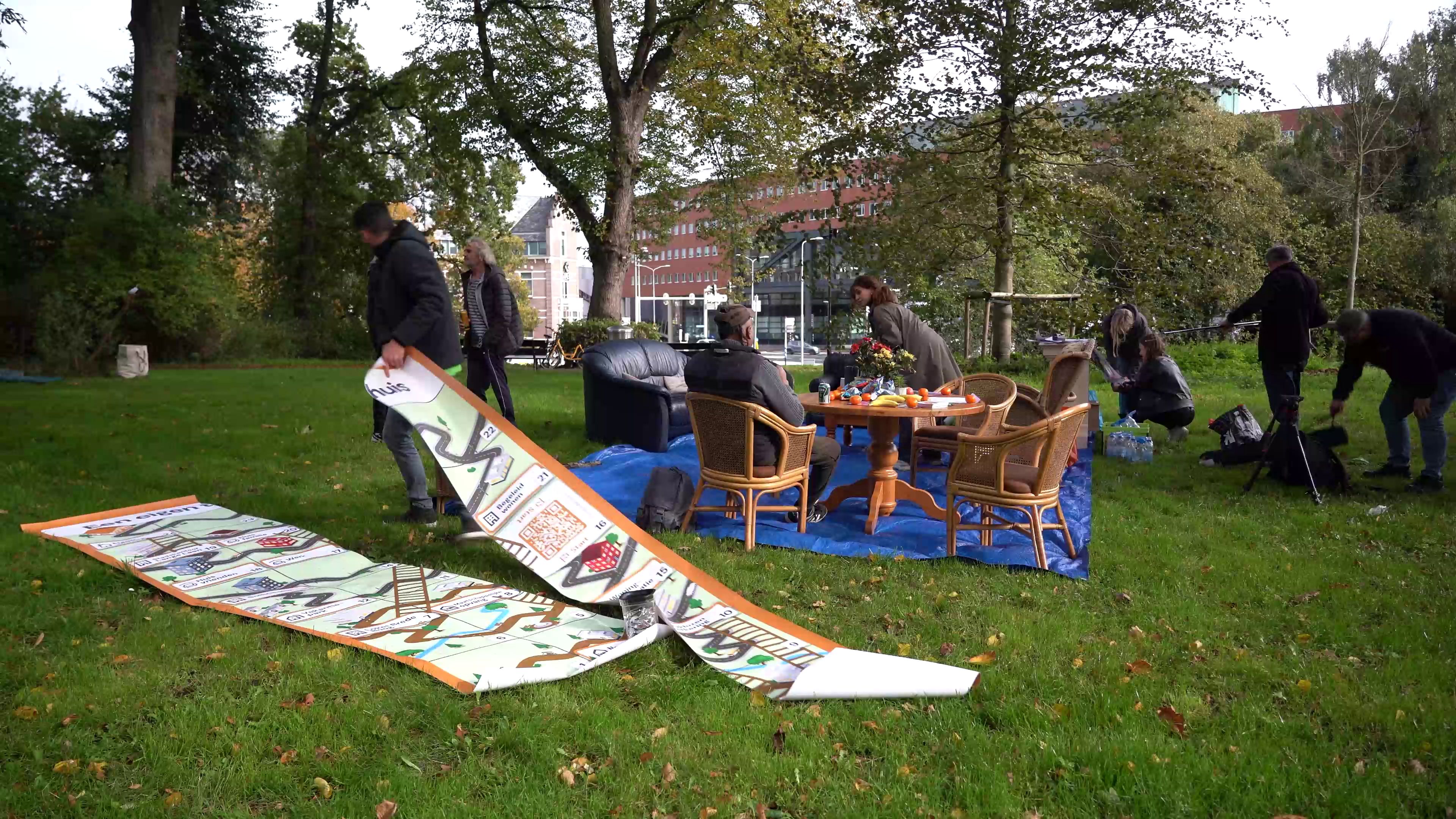 Mensen zetten een picknicksetting op in een park met een tafel, stoelen en een groot bord op de grond.