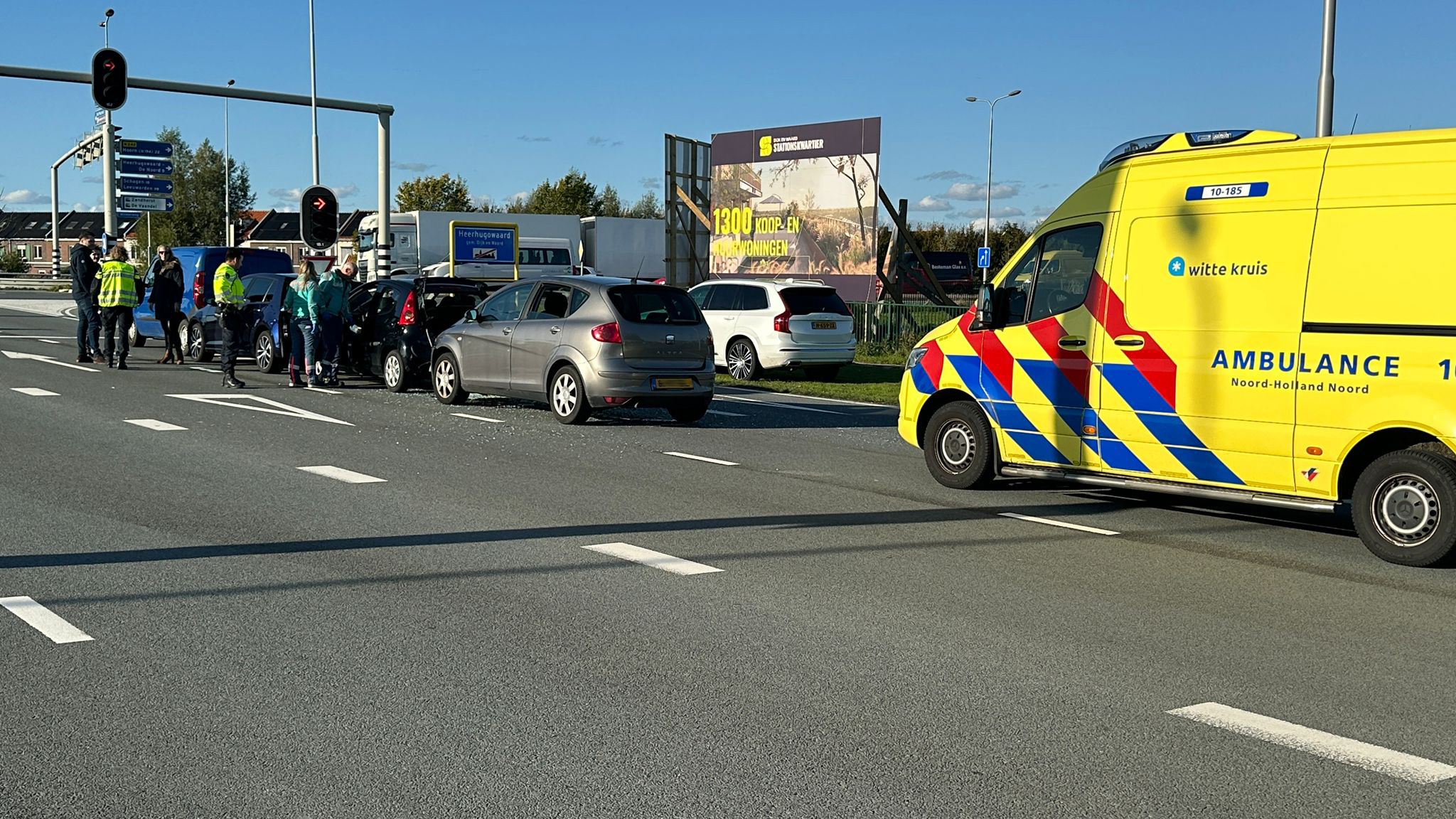 Verkeersongeluk op een kruispunt met meerdere auto's, omstanders, politieagenten en een ambulance aanwezig.