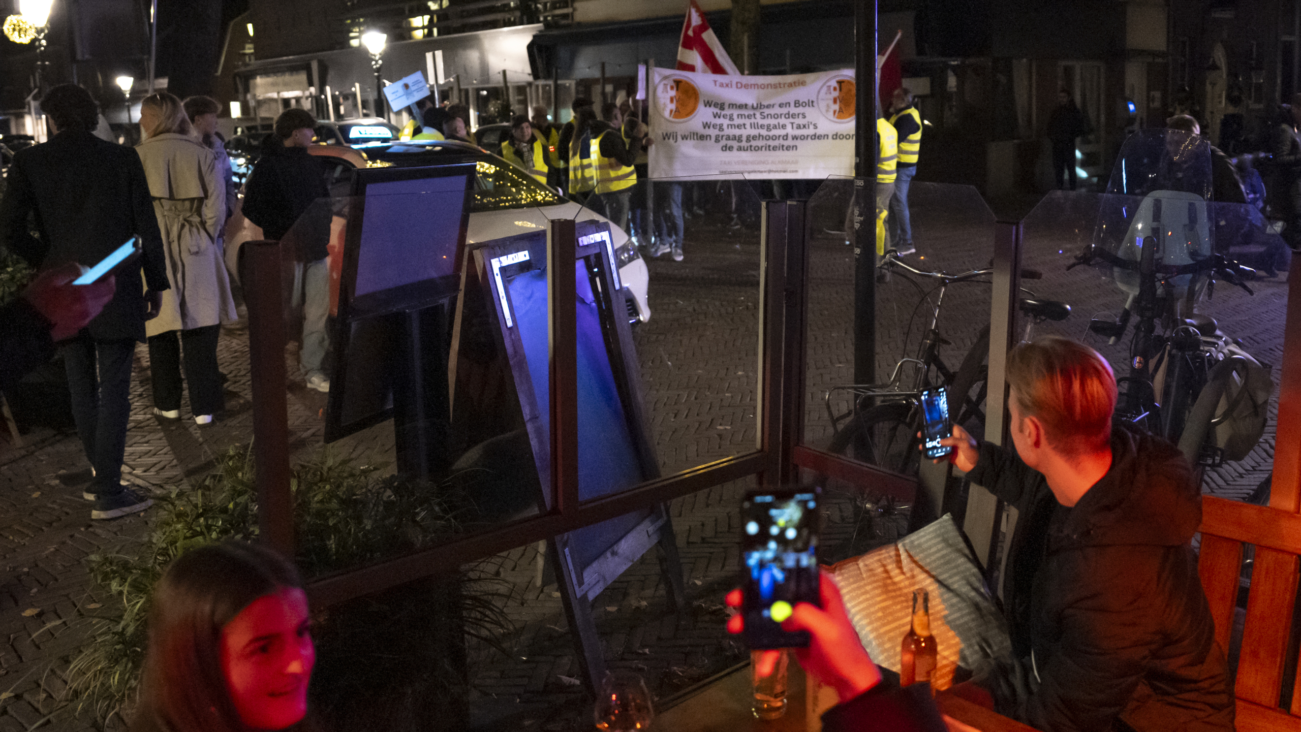 Protest met mensen in gele hesjes en een bord met tekst "Taxi Demonstratie" op straat, gefotografeerd vanuit een café.