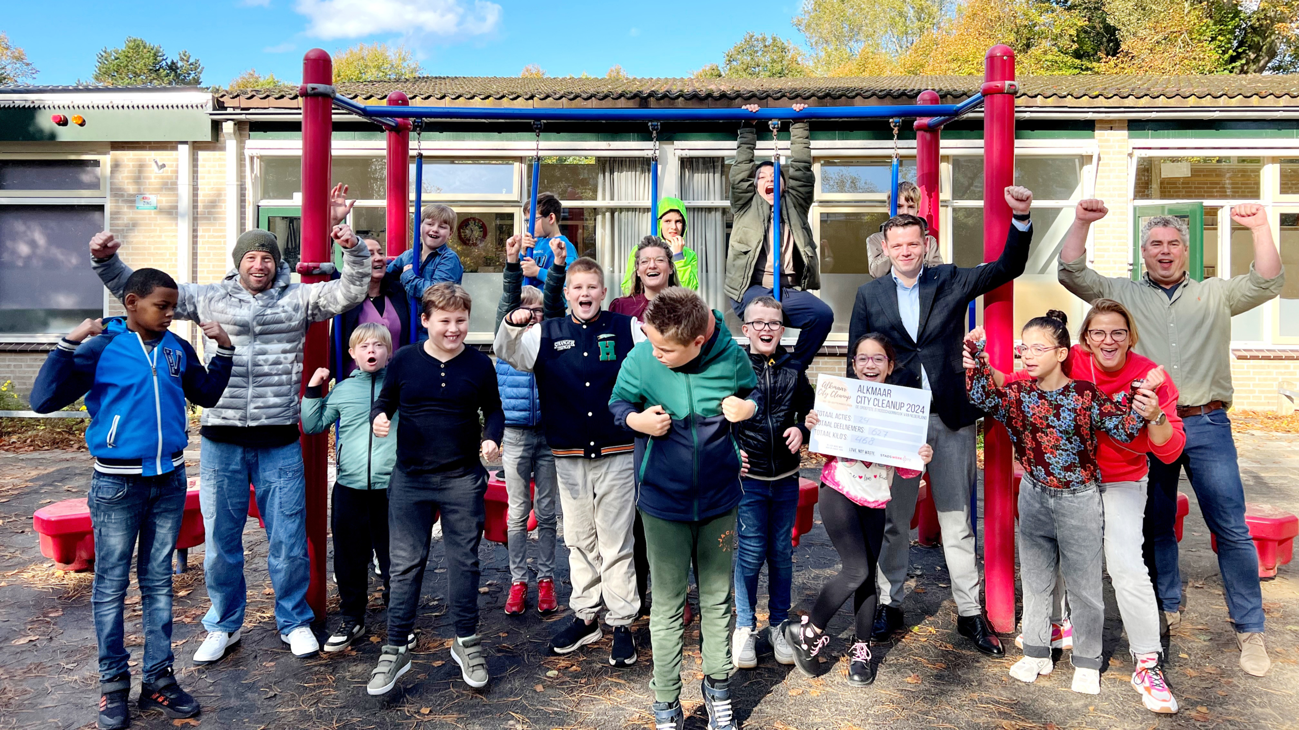 Een groep kinderen en volwassenen juichen op een speeltuin met een cheque voor de "Alkmaar City Cleanup 2024".